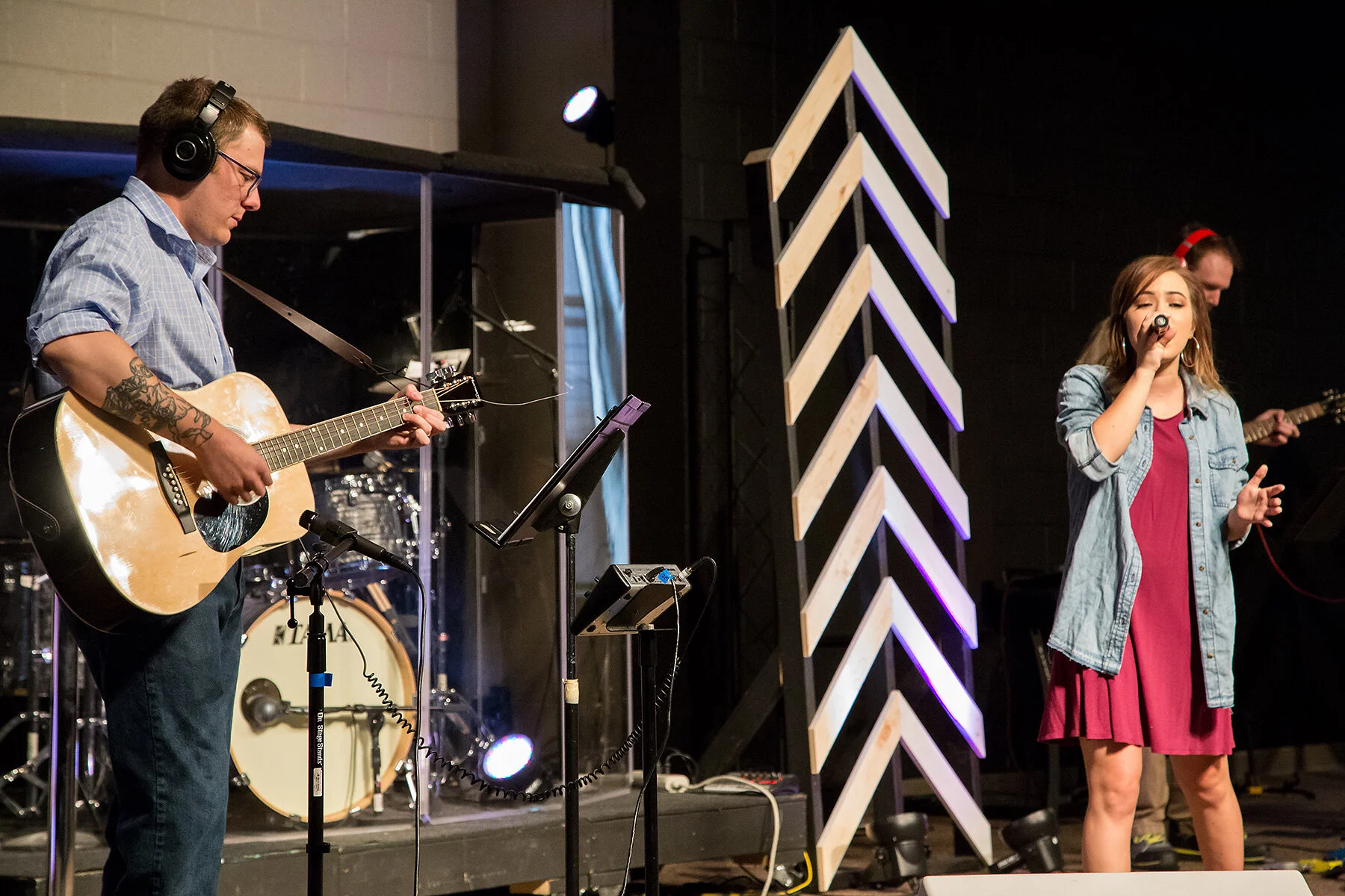 A band performing on stage with a male guitarist playing an acoustic guitar and a female singer holding a microphone, with other band members in the background.