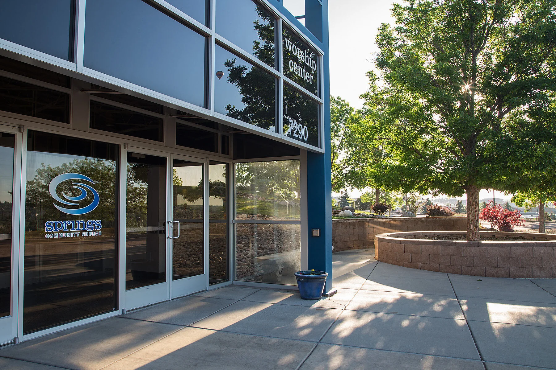 Exterior of Springs Community Church, with a glass entrance door featuring a blue and white logo, and a large window above with the words 'Worshio Center'. A sidewalk leads to the door, and there is a blue pot with plants next to the entrance. To the