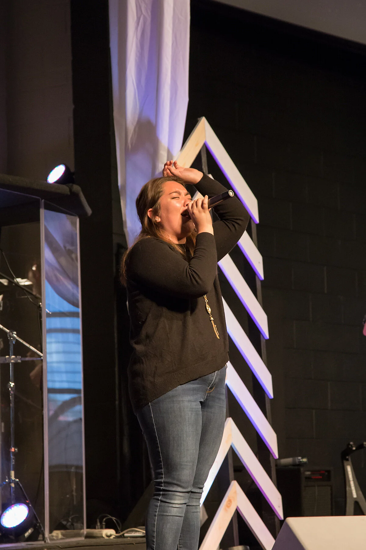 A woman singing into a microphone on a stage with black walls, stage lights, and decorative wood panels in the background.