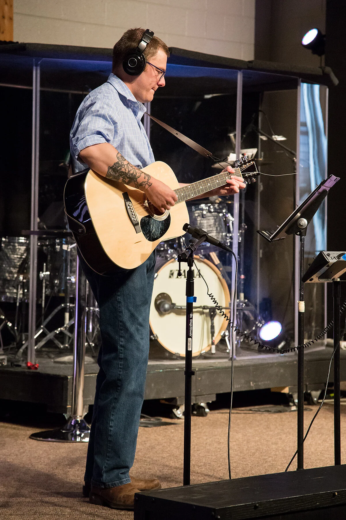 A man wearing glasses, headphones, a blue checkered shirt, and jeans is playing an acoustic guitar on stage in front of a microphone stand with music sheets. There is a drum set in the background behind a clear protective shield.