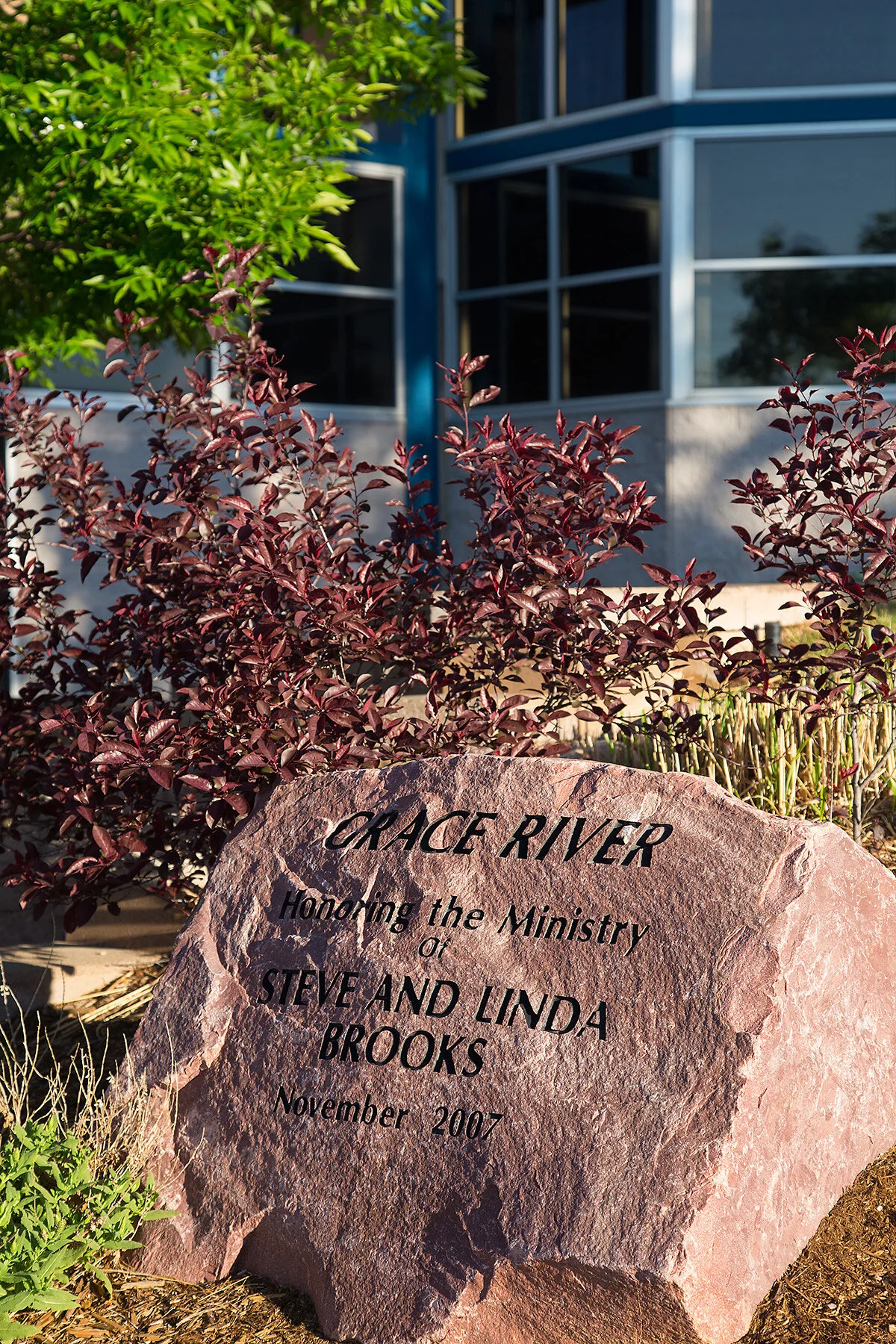 A large pinkish rock with engraved black text that reads, 'GRACE RIVER Honoring the Ministry of STEVE AND LINDA BROOKS November 2007' is surrounded by plants and bushes in front of a building with blue-gray siding and large windows.