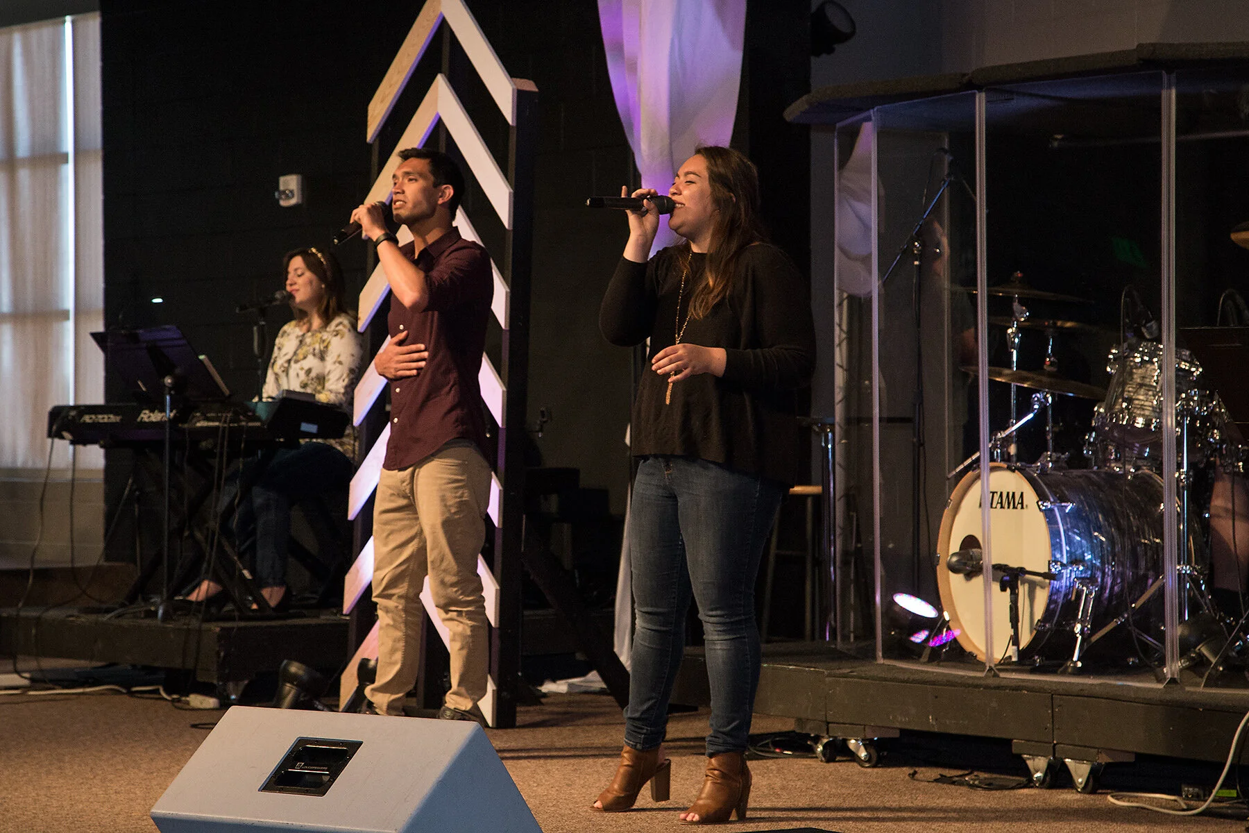 Three people perform on stage with a keyboard and drum set, two women and one man singing into microphones.