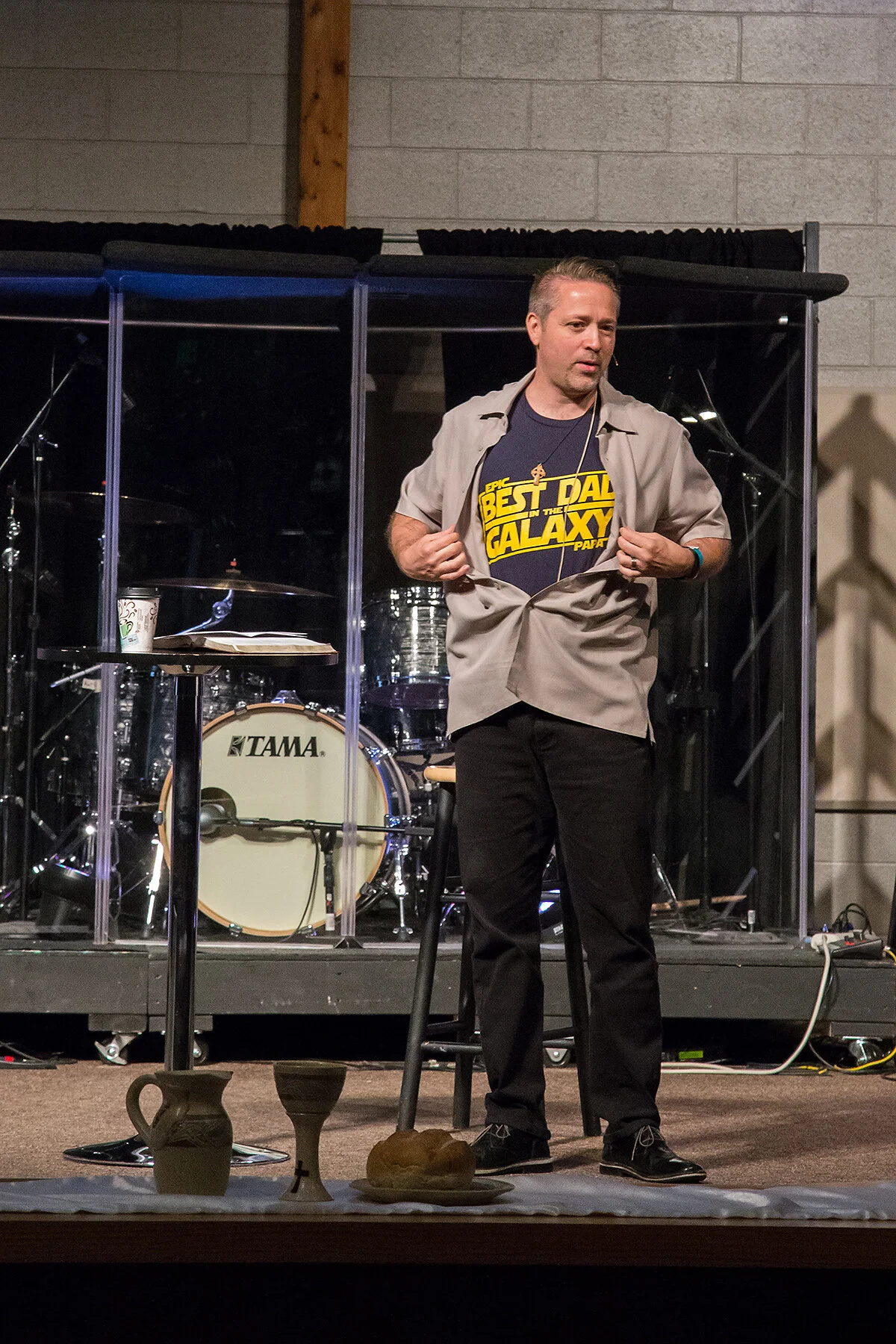 A man standing on stage in front of a drum kit, lifting his shirt to reveal a Star Wars themed T-shirt that says 'Best Dad in the Galaxy'. There are various items on a table in front of him, including a coffee cup, a plate with bread, and pitchers. T
