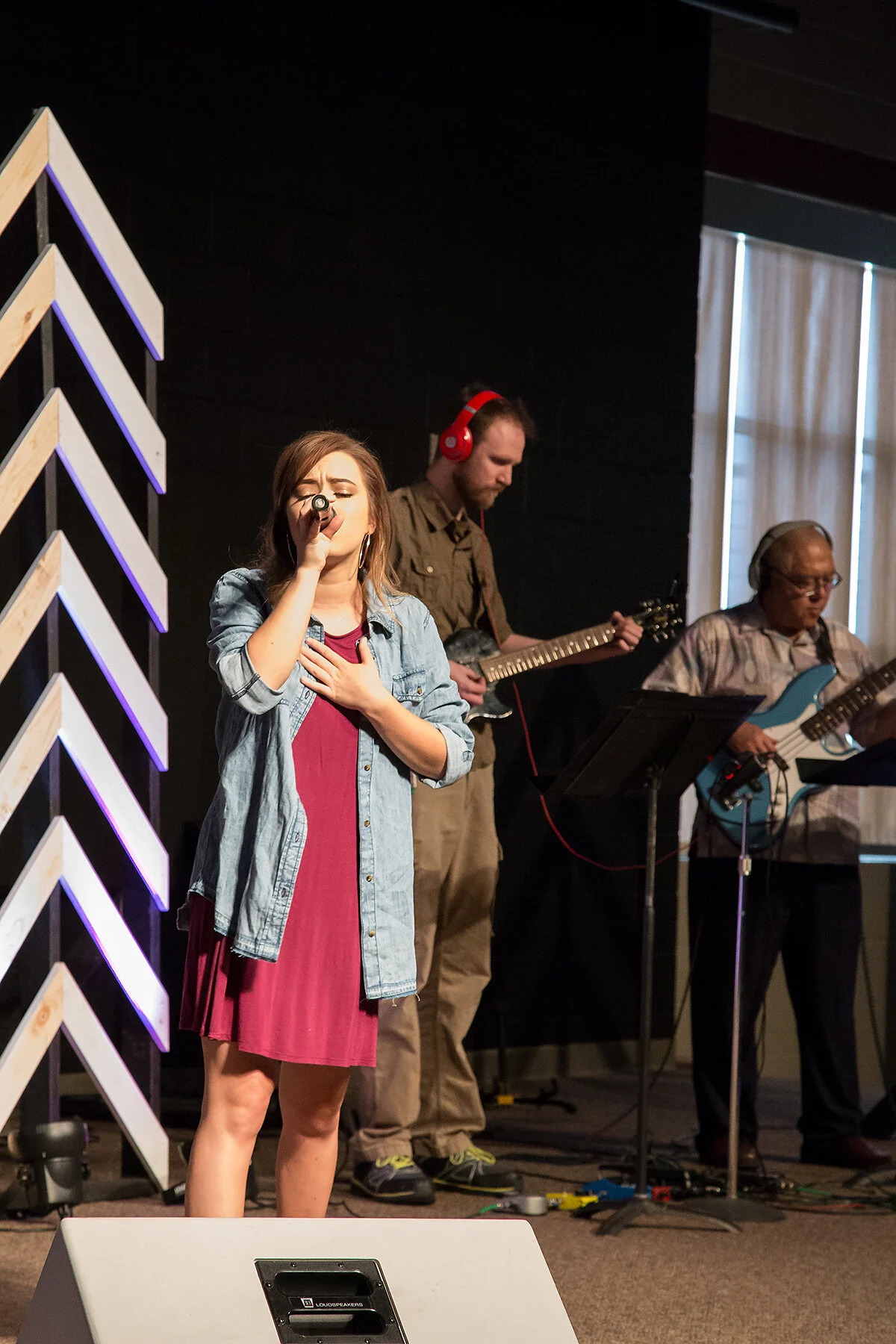 A young woman singing with her hand on her chest, wearing a denim jacket and a red dress, with two men playing guitars in the background, one with red headphones and the other with a bass guitar, on a stage with wooden paneling and vertical blinds.