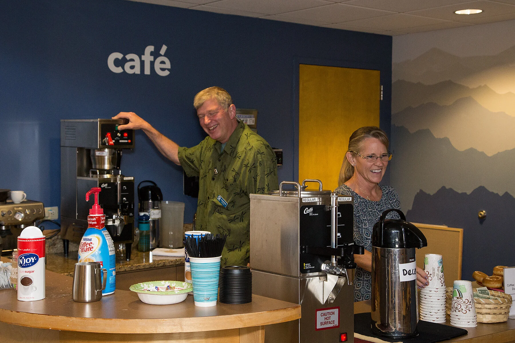 A man and woman smiling behind a coffee station at a cafe. The man is operating a coffee machine, and the woman is pouring from a large thermal coffee dispenser. The cafe sign is visible on the wall.
