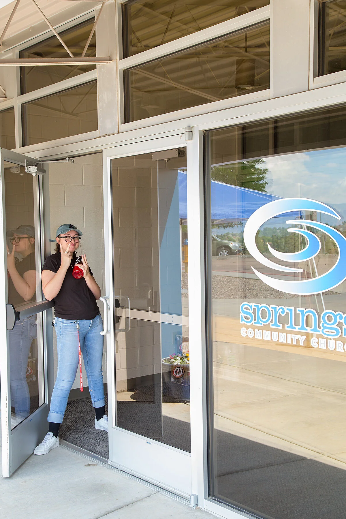 A woman standing outside the entrance of Spring Community Church, wearing glasses, a cap, black t-shirt, and blue jeans, holding a drink and making a peace sign with her fingers.