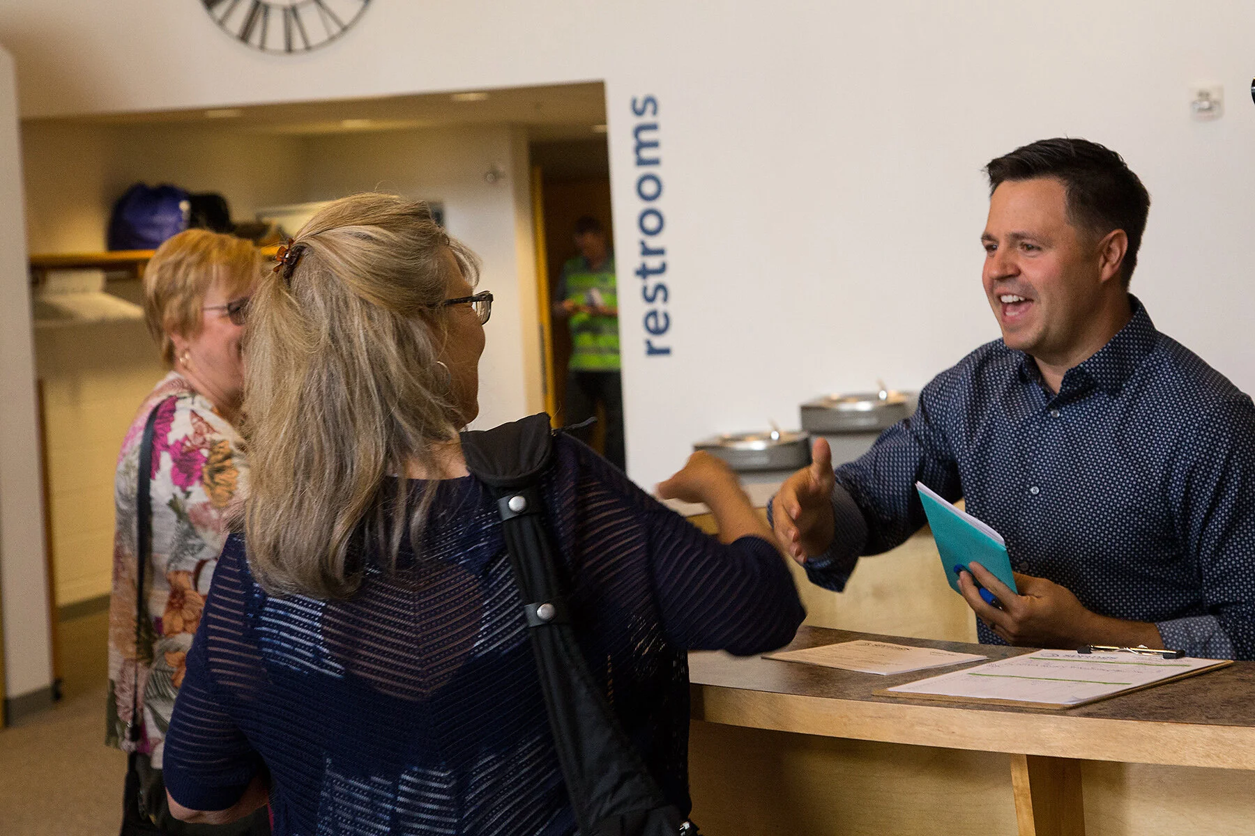 A smiling man at a reception desk greeting two women, one with glasses and blonde hair, in a wellness or medical center. The background features a sign that says 'restrooms' and a clock on the wall.