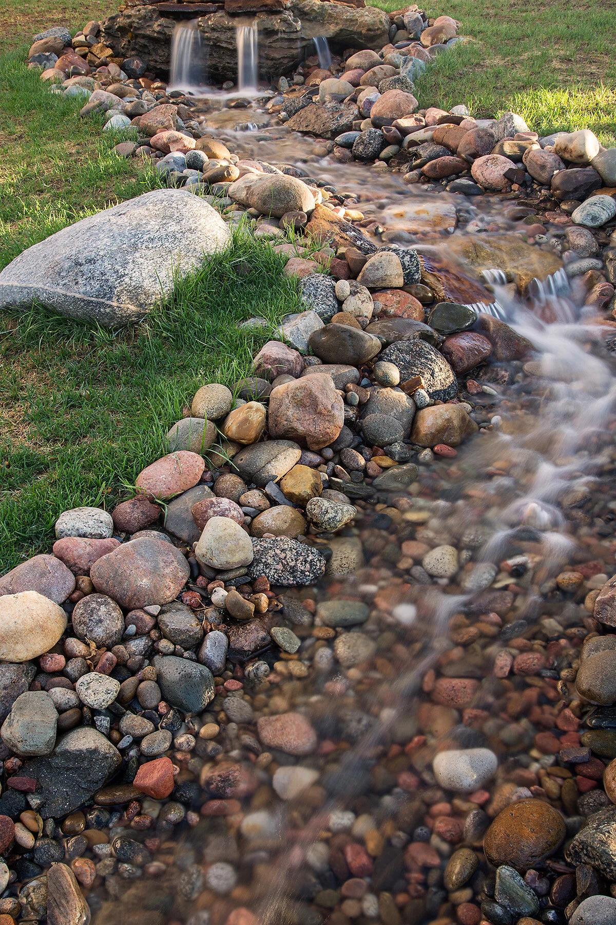 Small flowing stream with a rock-lined edge, flowing from a small waterfall surrounded by grass and multicolored rocks.