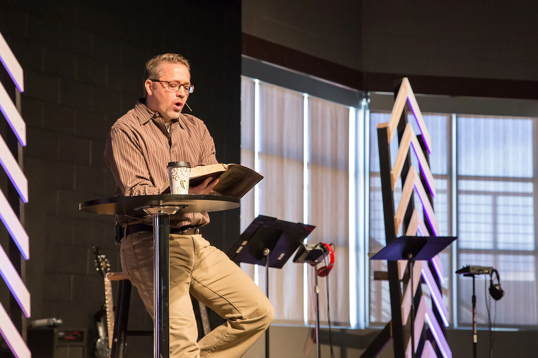 A man with glasses and a striped shirt reading from a book on a small round table, with a coffee cup nearby, on a stage with music stands and guitars.