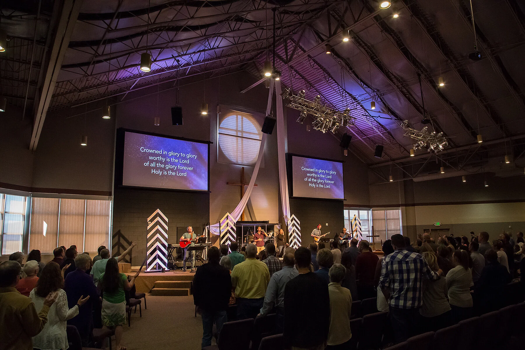 People gathered in a church auditorium, standing and singing during a worship service, with a band performing on stage, two large screens displaying lyrics, and a cross on the wall behind the stage.