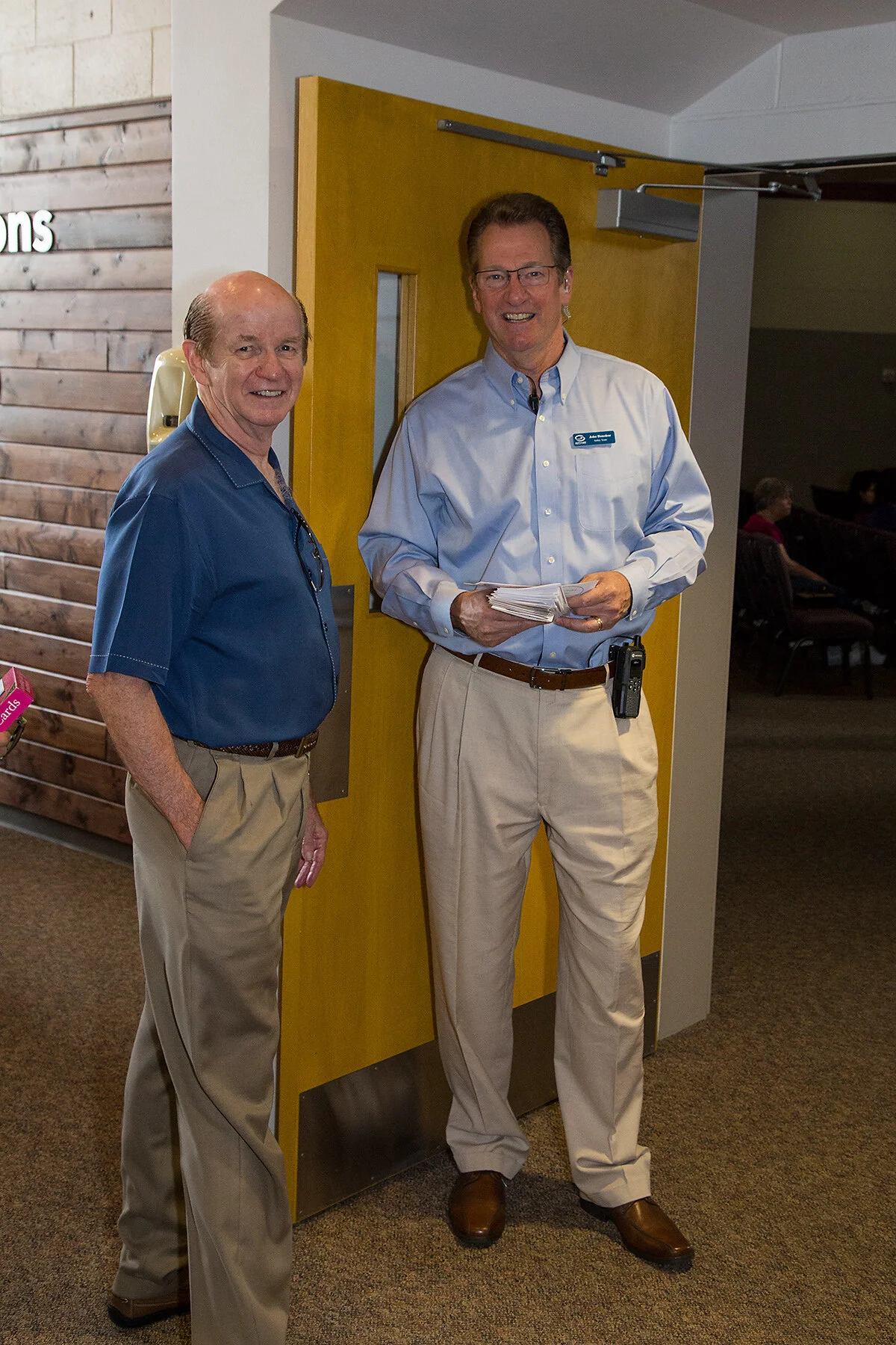 Two men standing indoors near a wooden wall and a yellow door. One man is older with a blue short-sleeve shirt and beige pants, smiling with his hand in his pocket. The other man is taller, wearing glasses, a light blue button-up shirt, beige pants, 