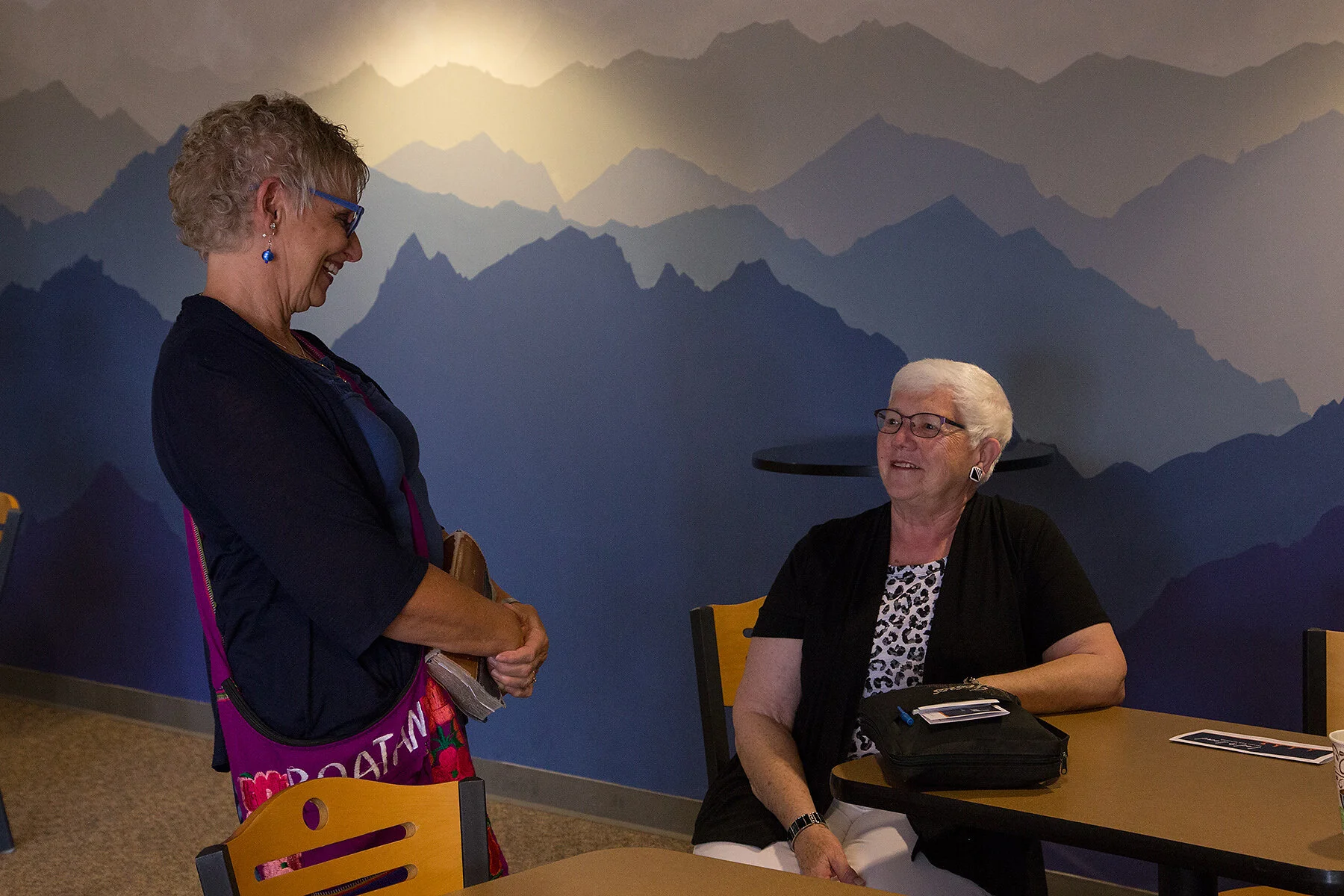 A woman standing and smiling, talking to another woman seated at a table, with a mountain mural in the background.