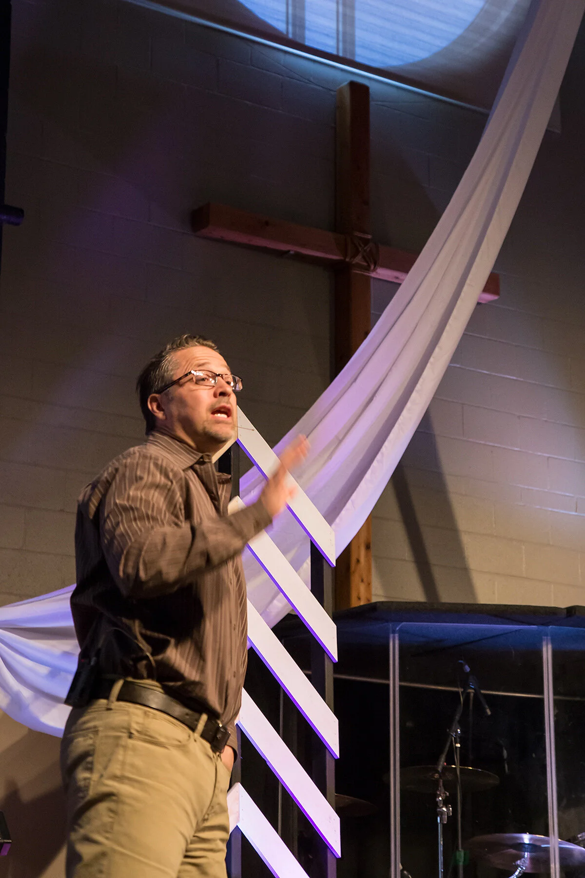 A man with glasses and wearing a striped shirt stands in front of a wooden cross and appears to be speaking or gesturing inside a church or religious setting.
