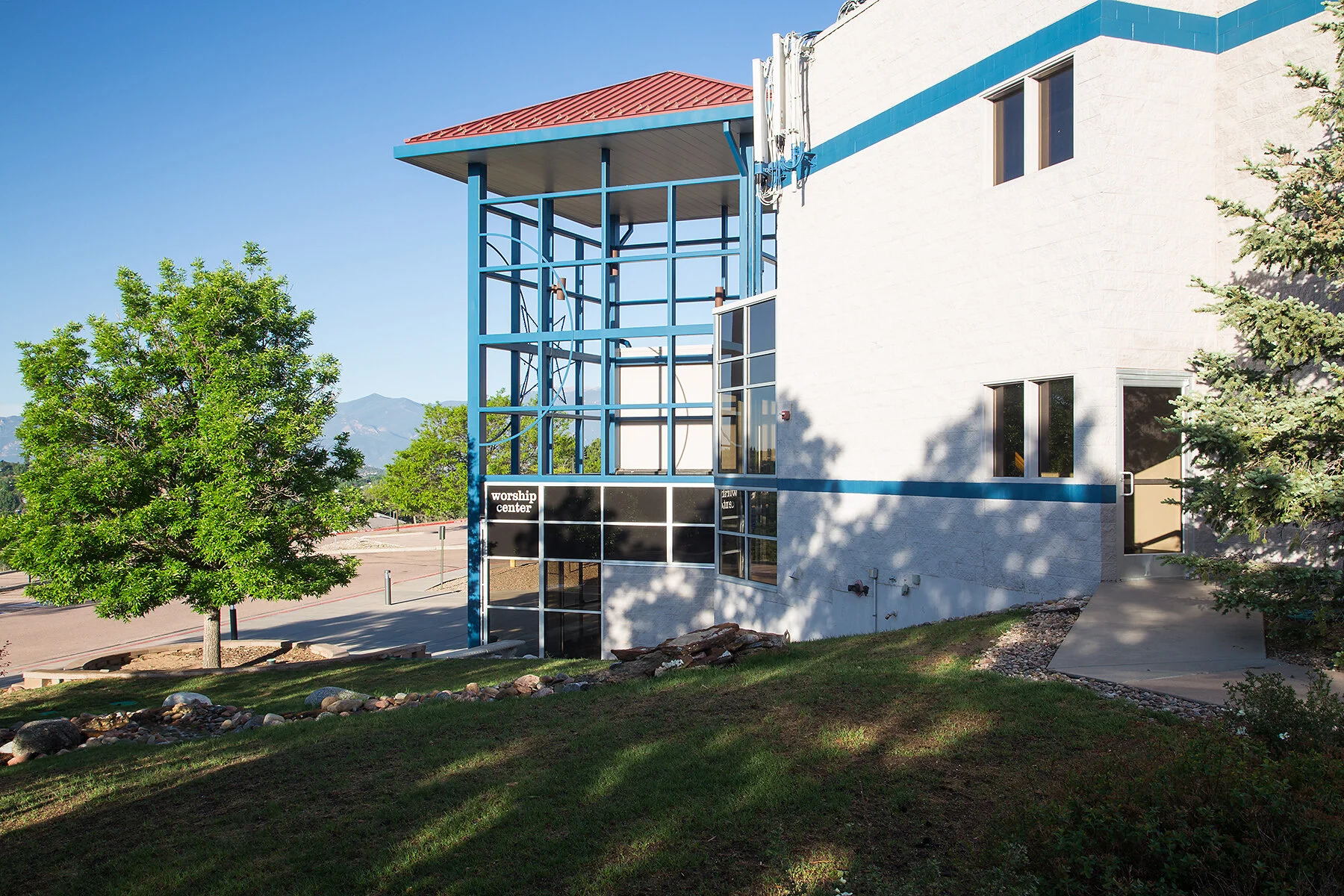 Exterior of a modern worship center building with a white facade, blue trim, and a red roof. There is a sign indicating the worship center on the front. The building features large windows and a sidewalk leading to the entrance, with trees and mounta