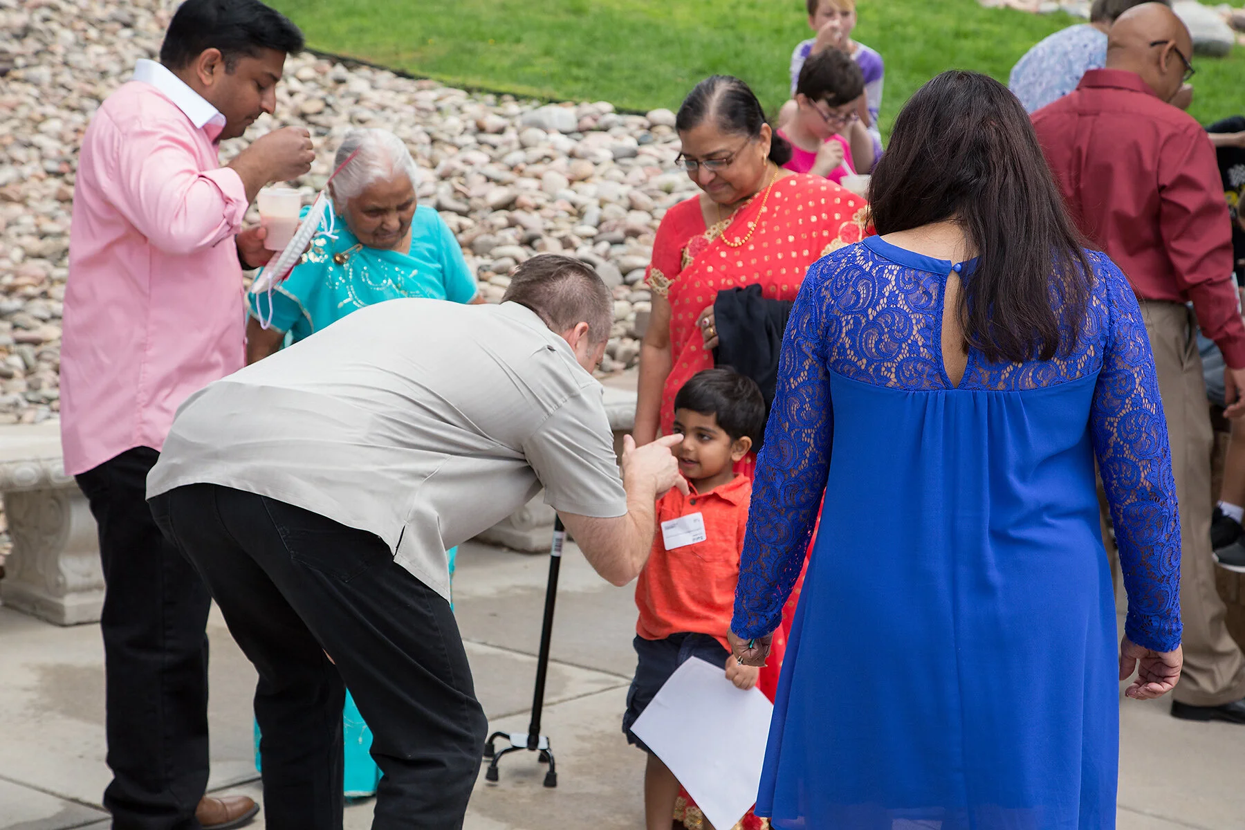 A group of people gathered outdoors, young and old, engaging in conversation and activities. A man kneels down to talk to a young boy who is holding a paper, while diverse women and children stand around them, dressed in colorful traditional and casu