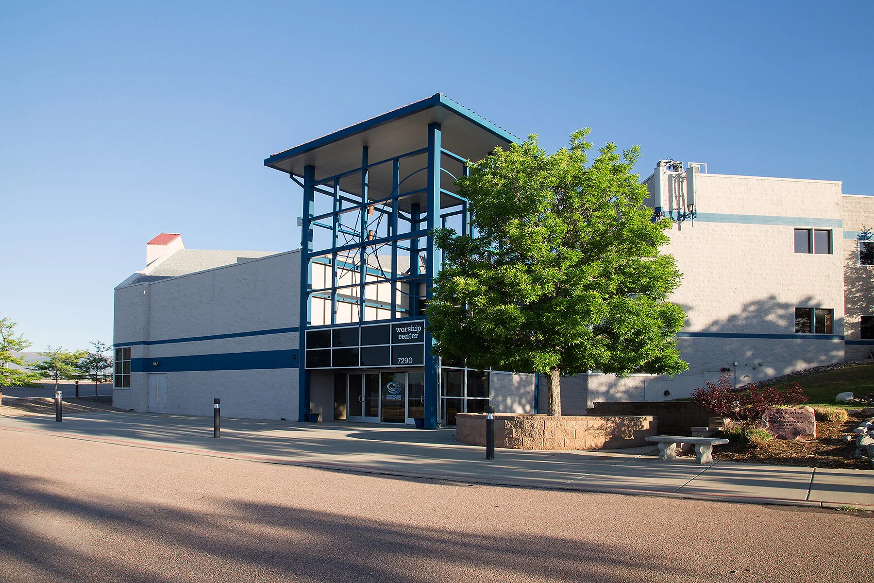 Modern white church building with blue accents and signage that reads 'worship center' at 7290, with a prominent green tree in front, a bench and flower bed nearby, and a clear blue sky overhead.