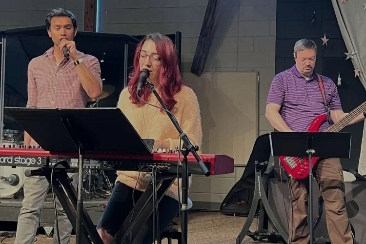 Three musicians performing on stage: a woman singing and playing keyboard, a man singing into a microphone, and a man playing electric guitar.