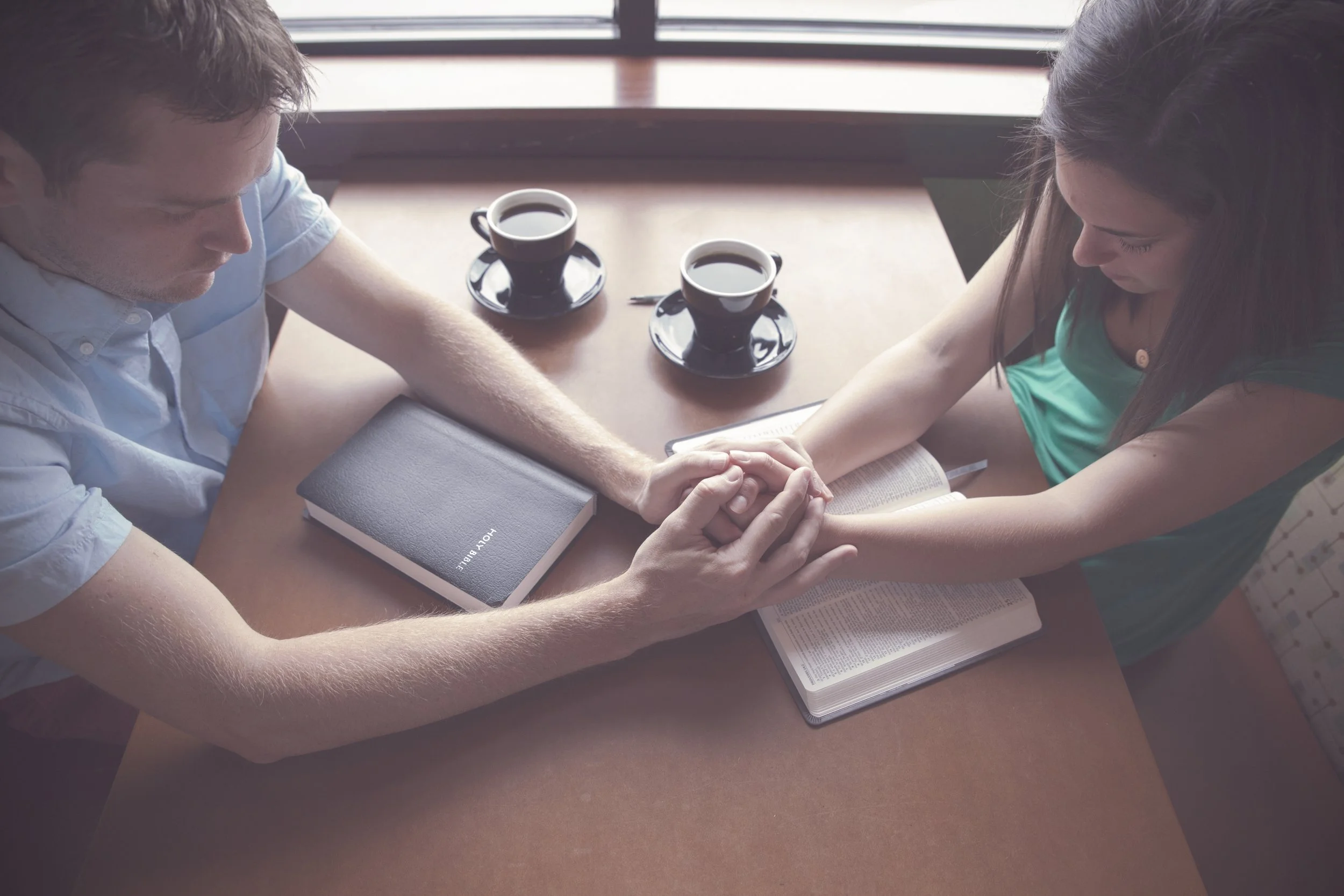 A man and a woman sit across from each other at a table, holding hands with their arms extended, over a Bible and an open book. Two cups of coffee are on the table.