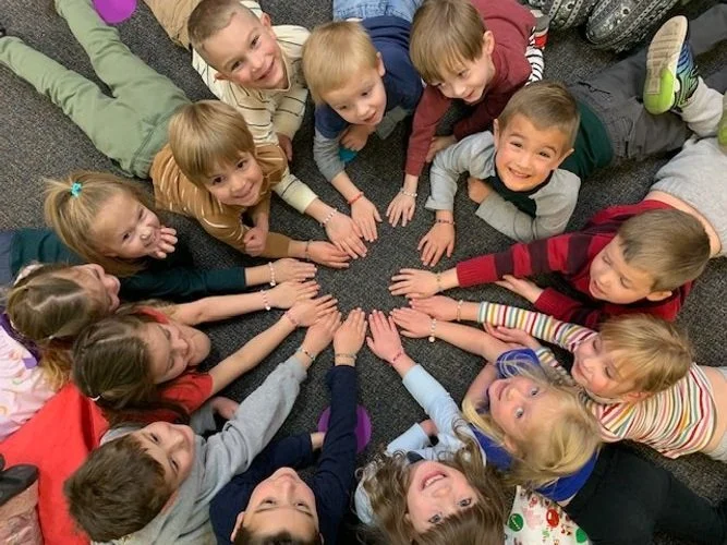 Group of children lying on the floor in a circle, reaching towards the center with their hands, smiling and looking up.