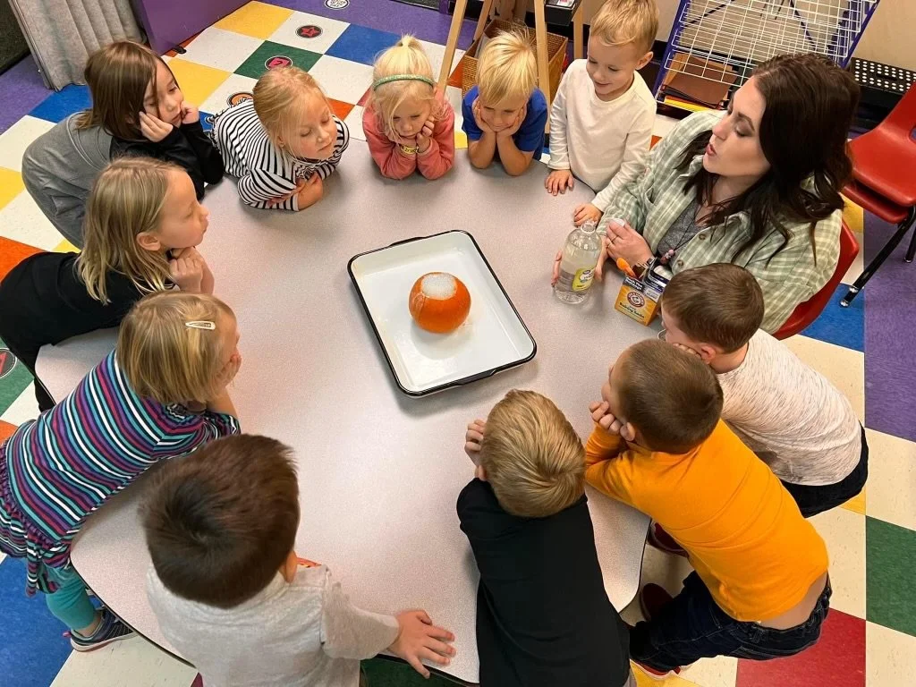 A group of young children sitting around a table, watching a science demonstration involving a pumpkin on a tray with liquid nitrogen, creating fog or smoke.