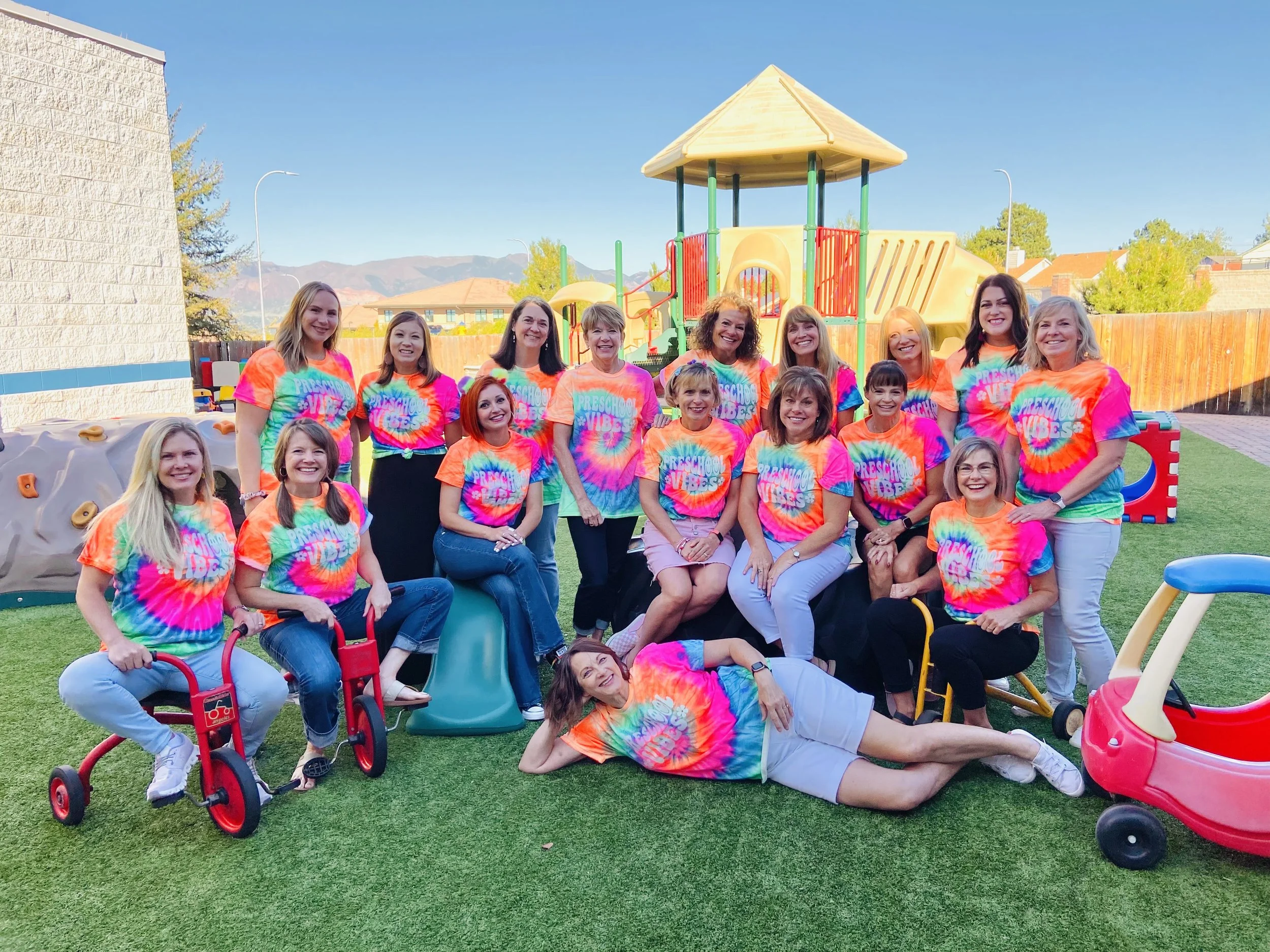 Group of women and girls in colorful tie-dye shirts at a playground, smiling for the camera on a sunny day.