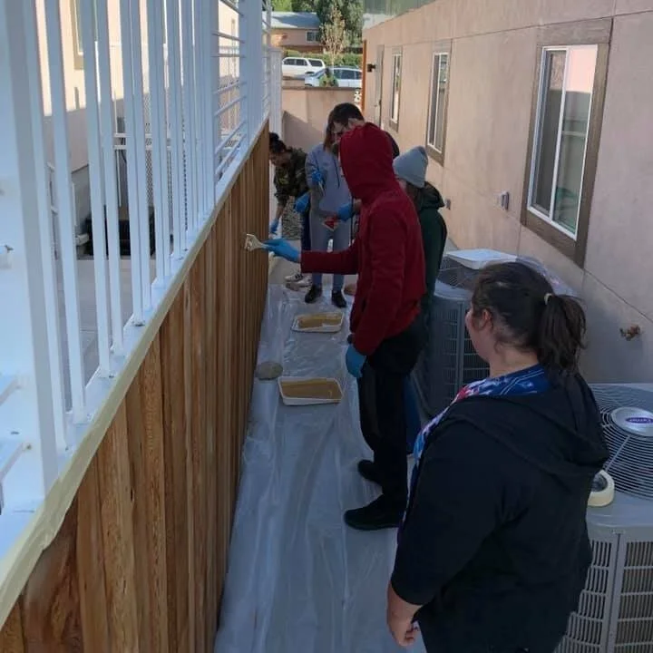 People standing in a line painting a wooden fence on a patio, using brushes and rollers, with trays of paint.