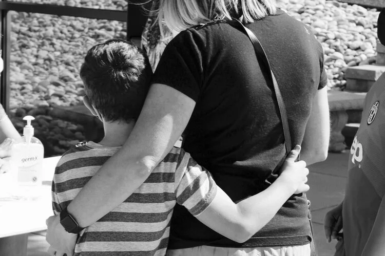 A person with a child, seen from behind, outdoors, with a table of hand sanitizer bottles nearby.