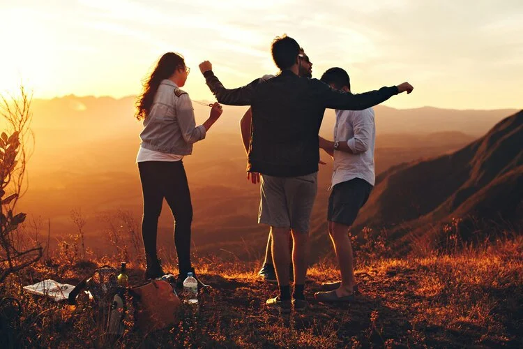 Four friends enjoying a sunset hike with scenic mountains in the background, standing on a grassy hill.