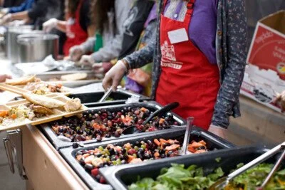 People serving and preparing food at a buffet or food station, with various dishes and salads in black containers.