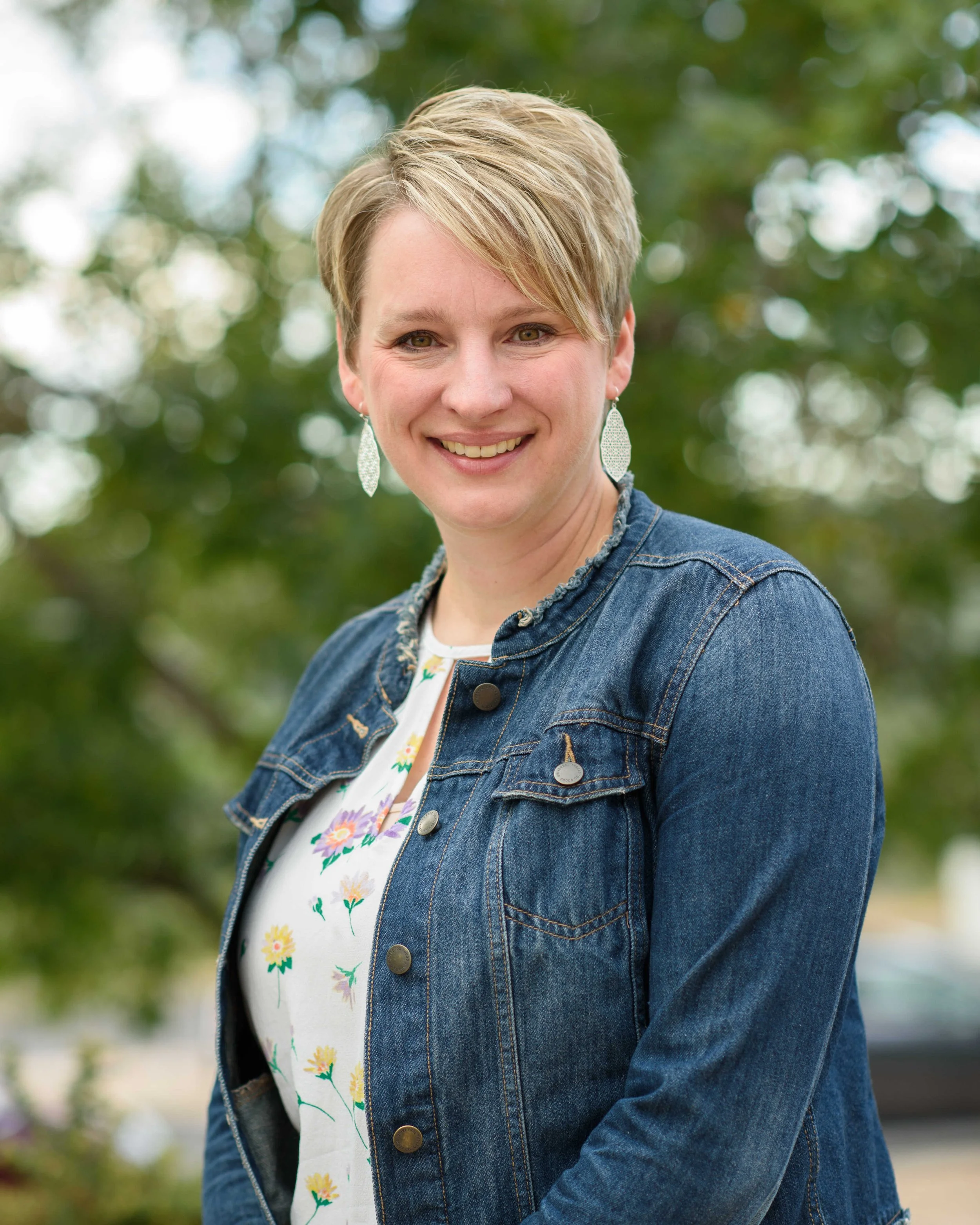 A woman with short blonde hair, wearing a denim jacket and patterned blouse, smiling outdoors with trees in the background.