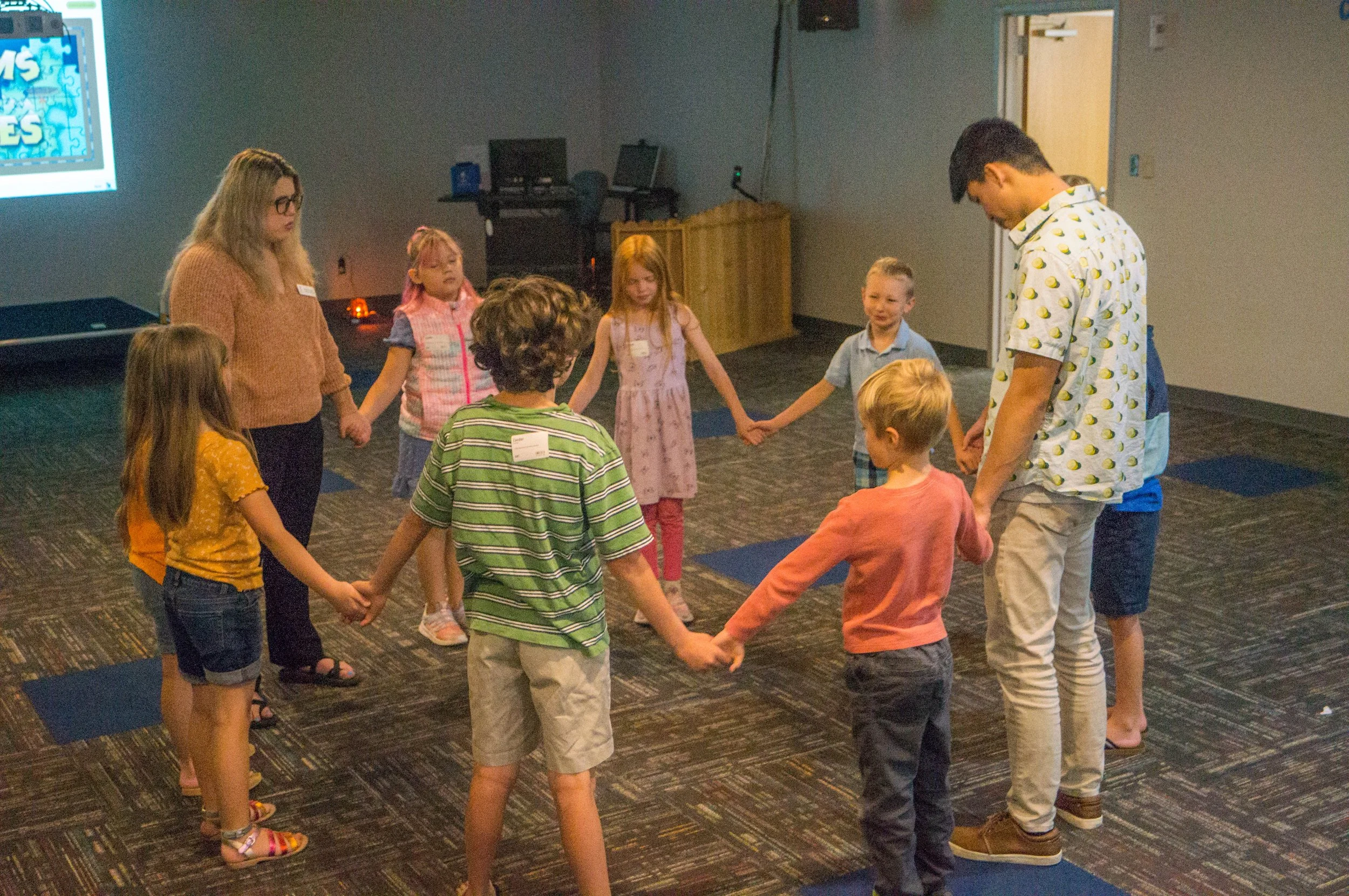 Children and adults holding hands in a circle in a room with a screen displaying puzzle pieces.