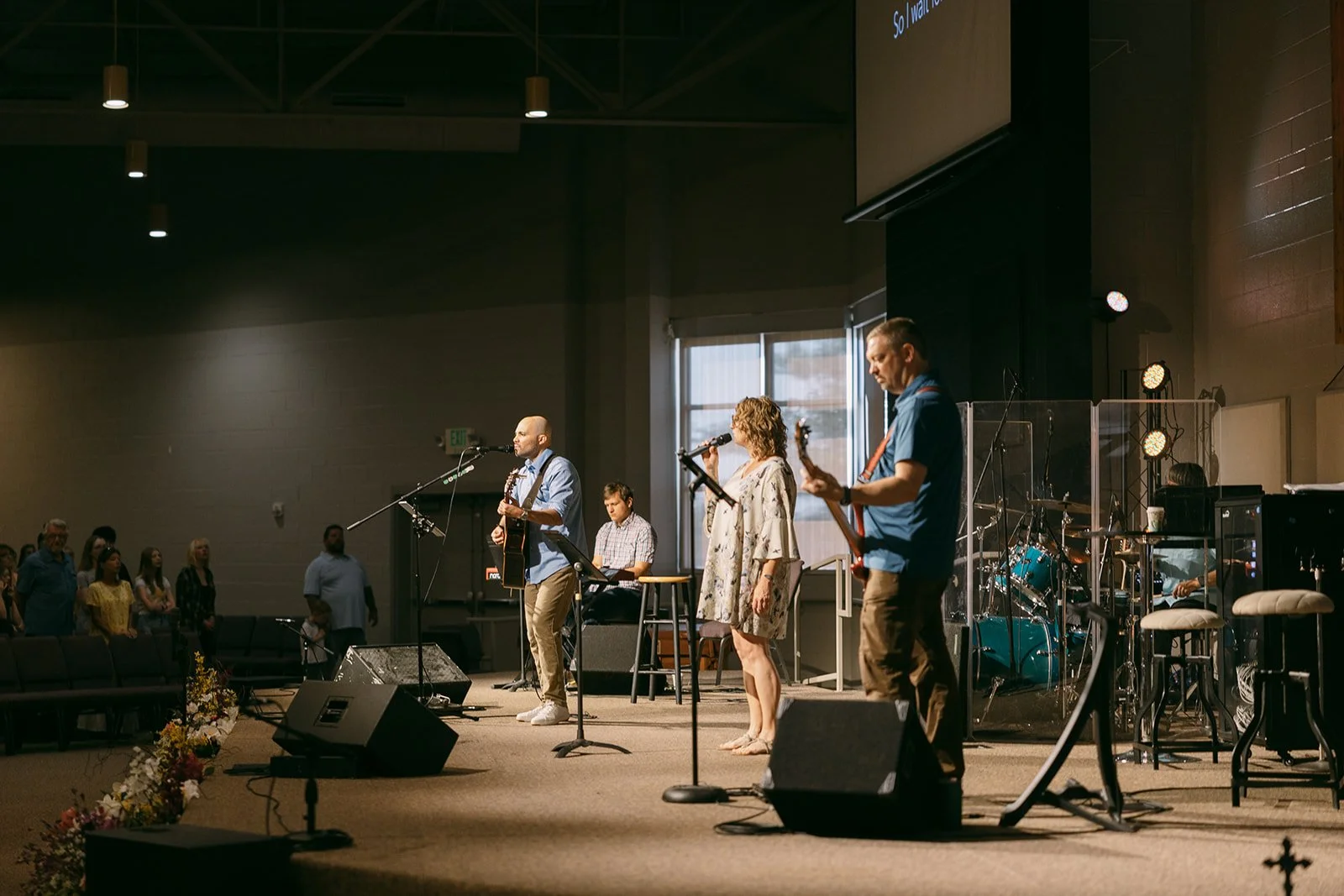 Three musicians performing on stage: a woman singing and playing keyboard, a man singing into a microphone, and a man playing electric guitar.