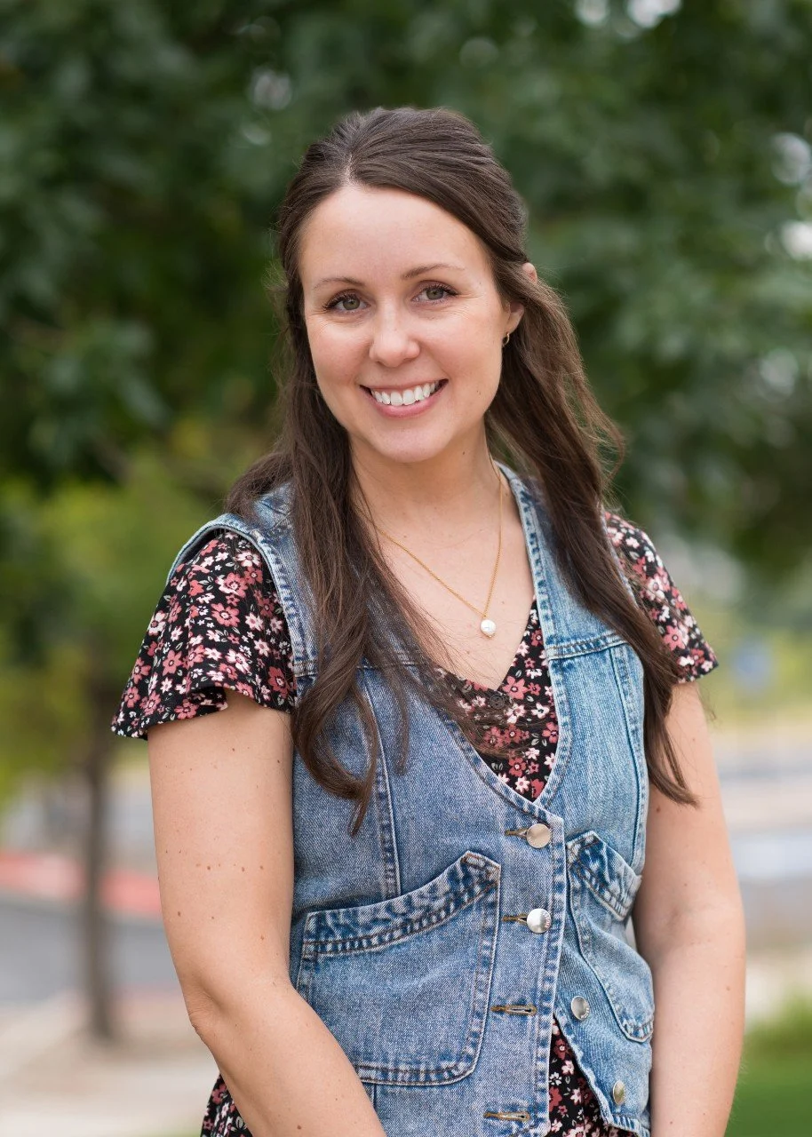 Young woman smiling outdoors, wearing a denim vest over a floral top, with long brown hair and a pearl necklace, with green trees in the background.