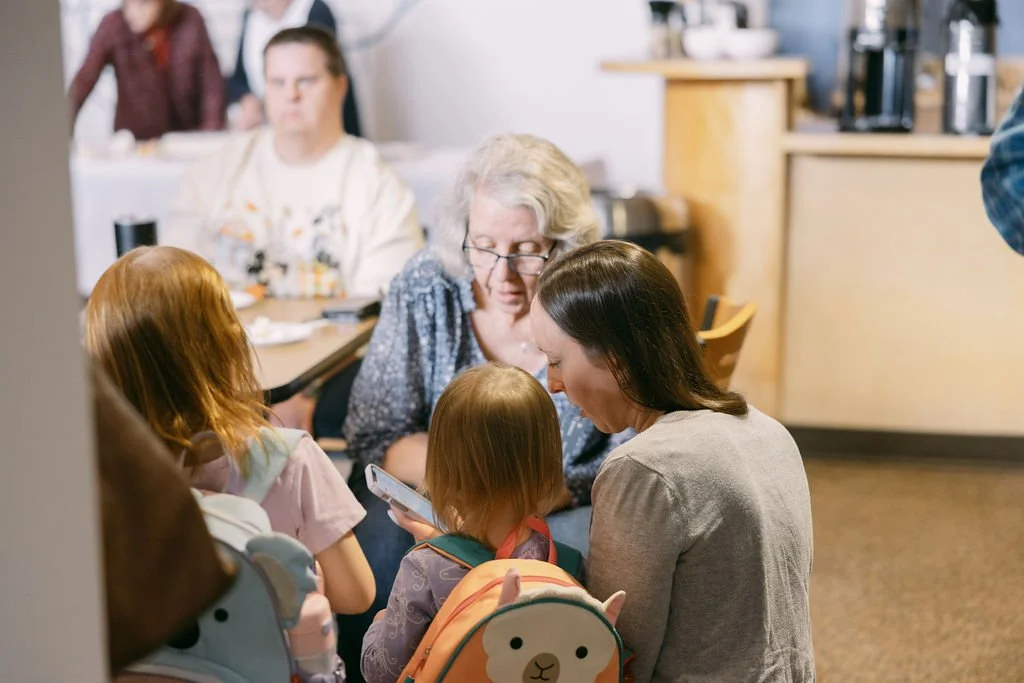 A group of children and adults gathered around a woman looking at a smartphone inside a cafe or restaurant.