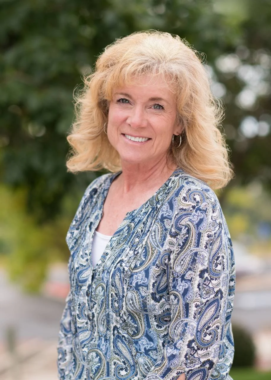 A smiling middle-aged woman with blonde, wavy hair outdoors in front of green trees, wearing a patterned blue and white blouse.