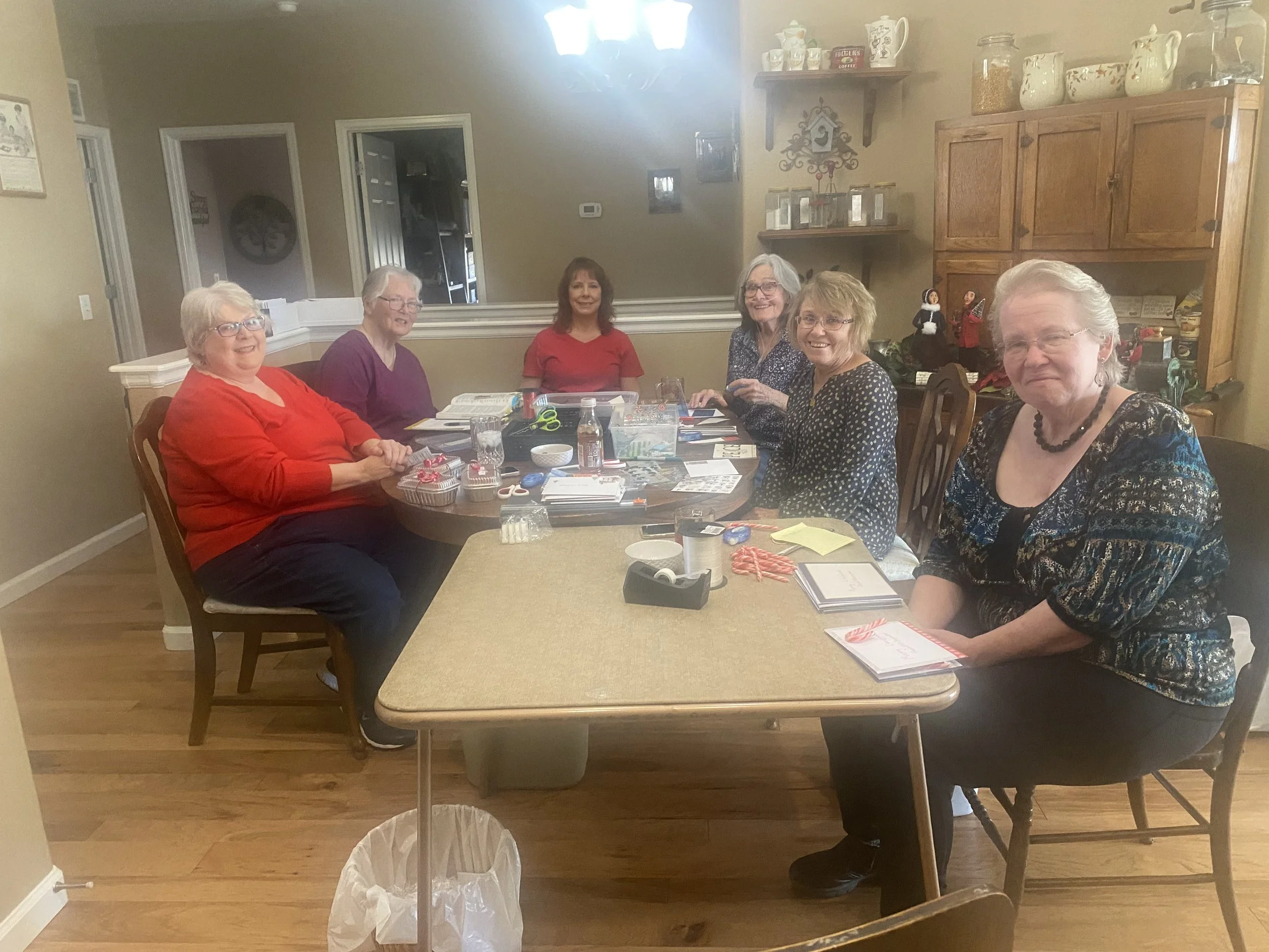 A young boy in a maroon shirt reaching for pineapples on a store shelf, holding a shopping bag and a plastic container. A young girl is nearby, holding a large purple bag and a plastic container.