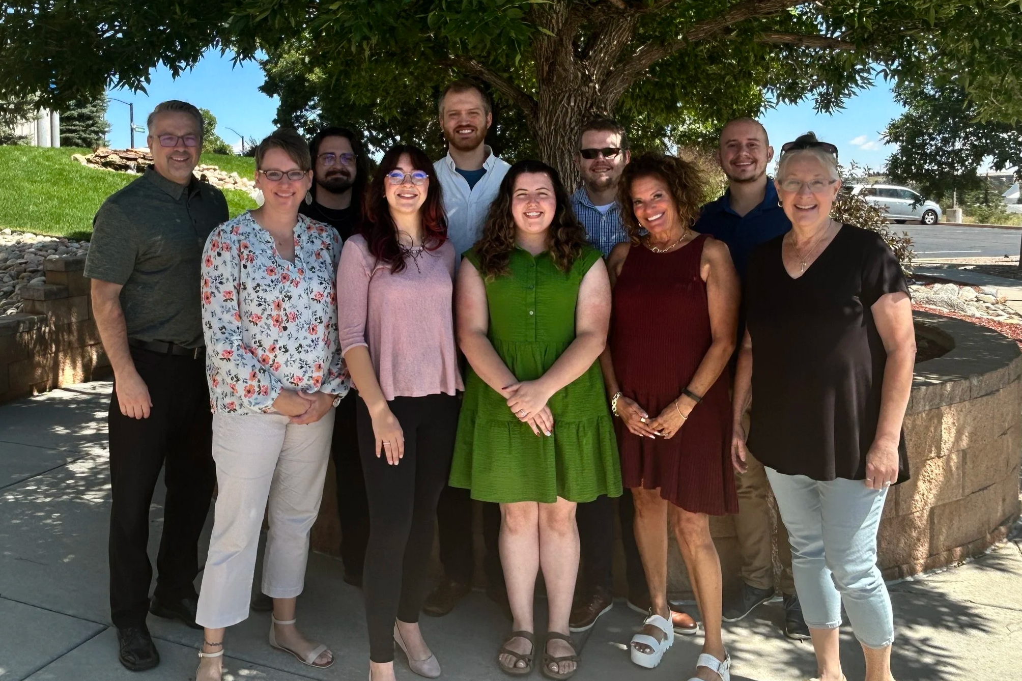 A group of eleven diverse people standing outdoors under a large green tree, smiling, with a stone planter and a parking lot in the background.