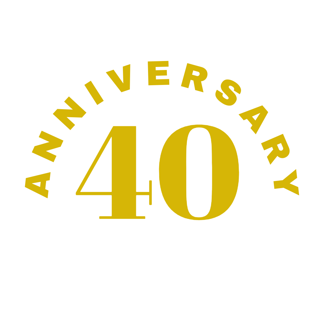 A man with curly hair and glasses smiling while sitting at an audio mixing console in a dimly lit studio or auditorium.