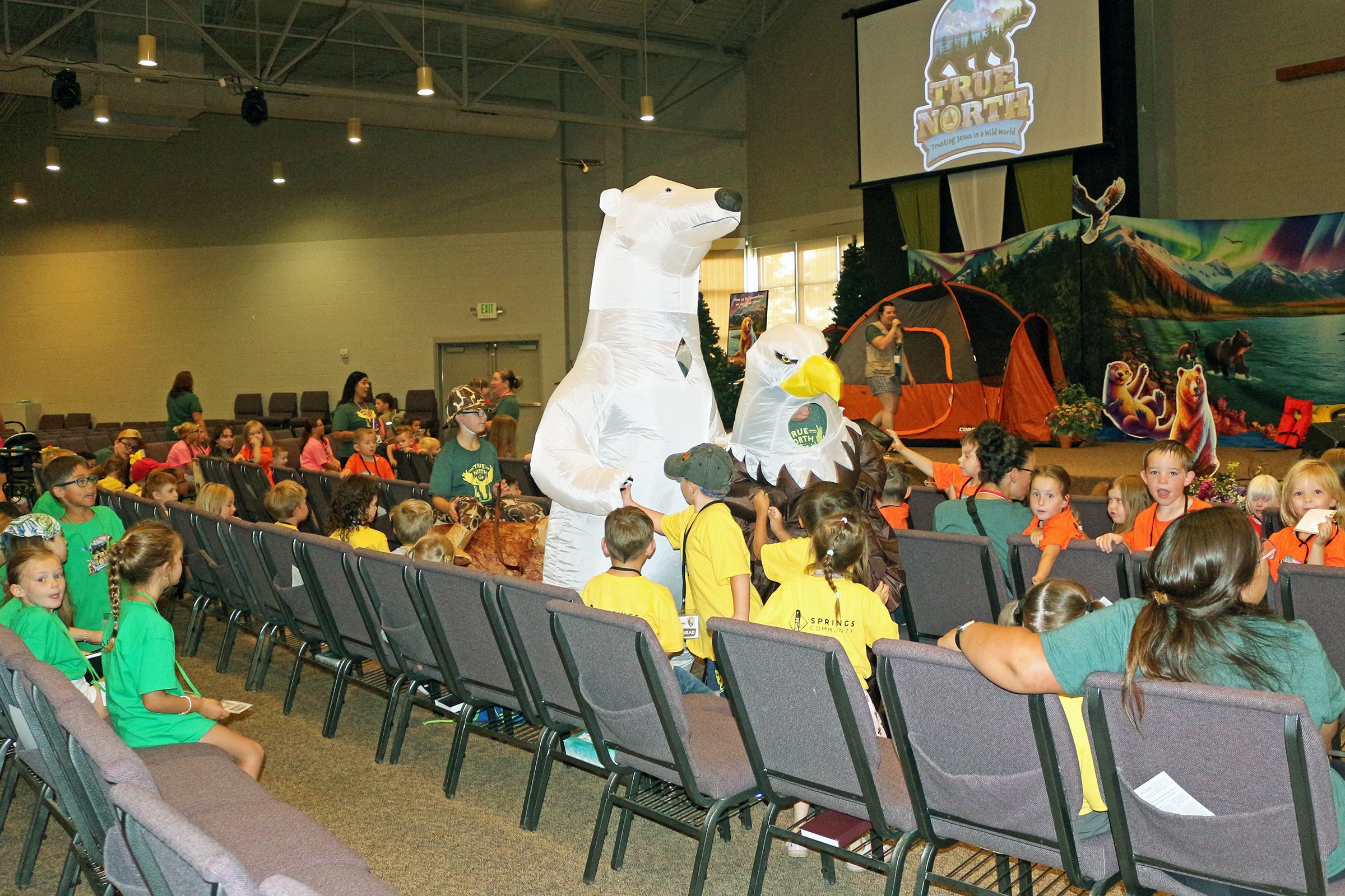 Children sitting on the floor watching a stage performance at an indoor event with a marine life theme called "SCUBA" and colorful ocean-themed decorations.