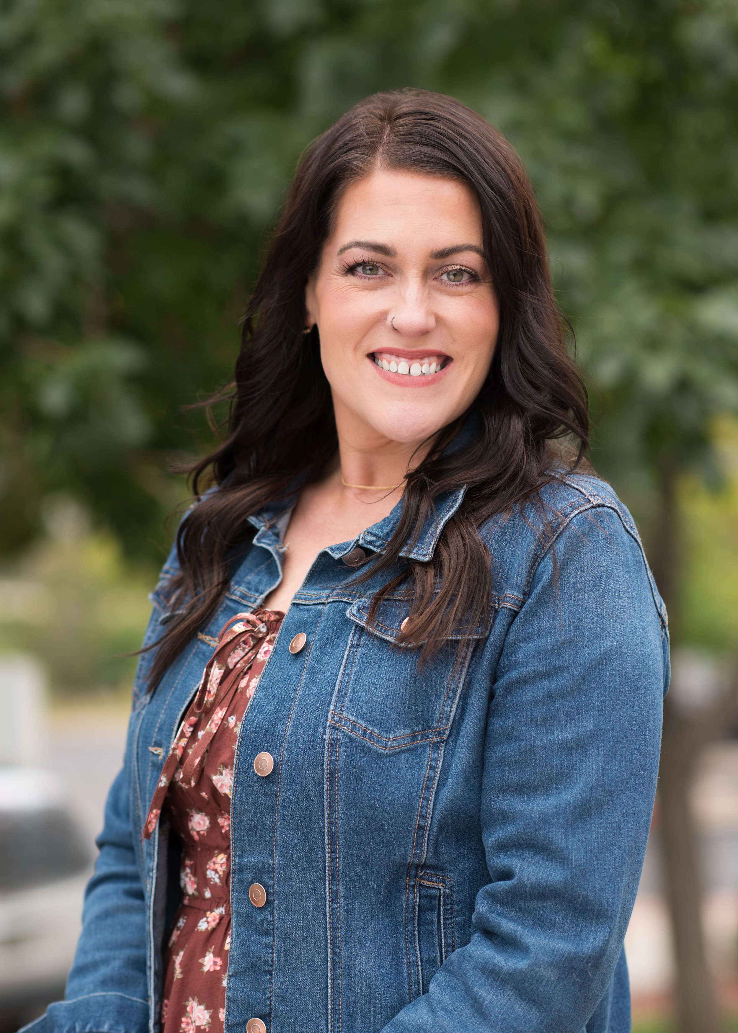A woman with dark brown hair, wearing a denim jacket over a red floral dress, smiling outdoors with blurred green trees in the background.