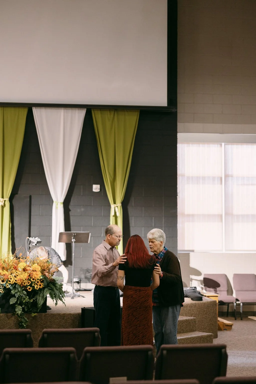 Three people engaged in conversation on a stage in a church or auditorium, with a woman with red hair in the center and two men, one with gray hair and the other with glasses. The stage has flowers and curtains in the background, with seating in front.