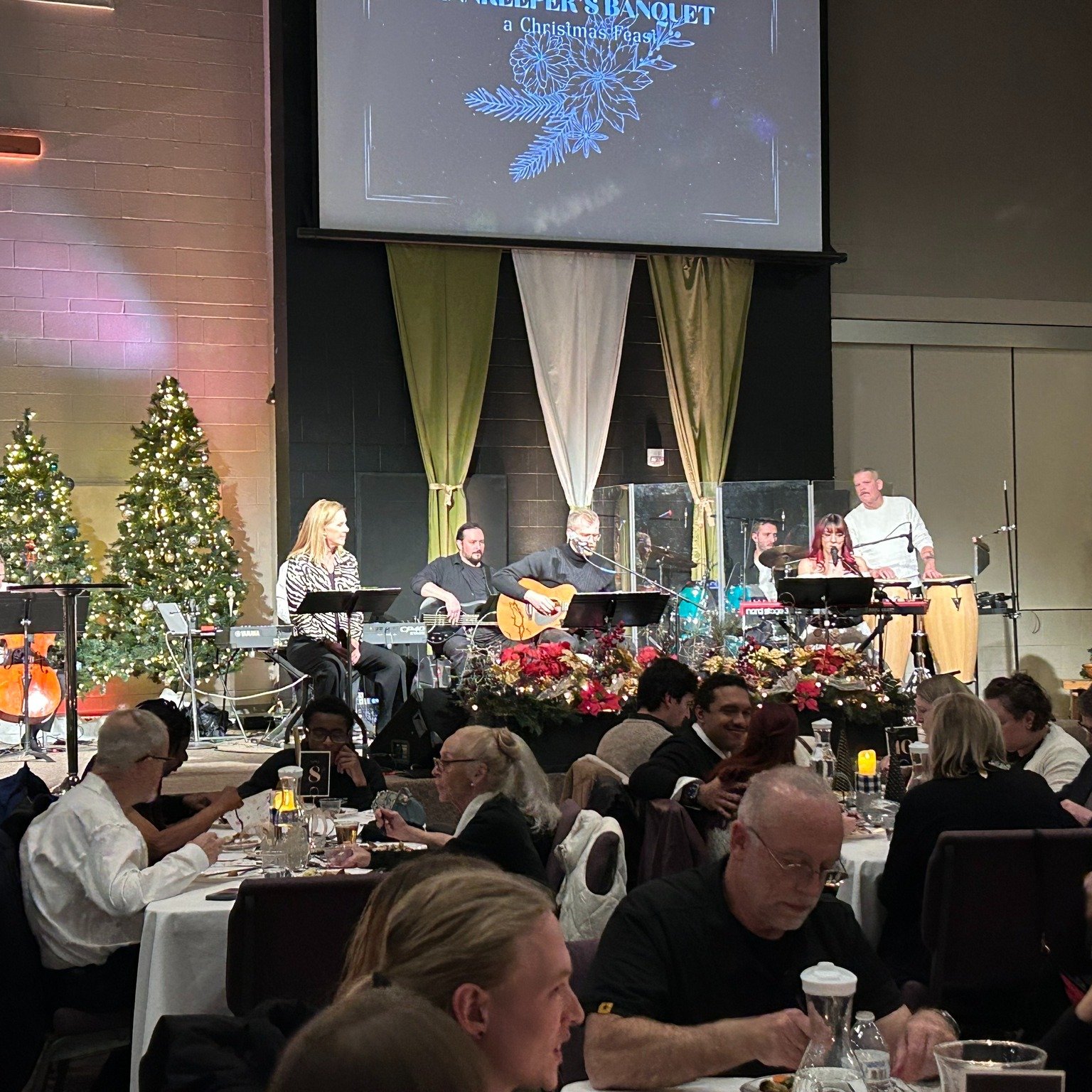 Children and adults seated at a long banquet table decorated with holiday-themed items during a festive event, with a stage in the background featuring Christmas trees, musical instruments, and music equipment, under holiday lighting.