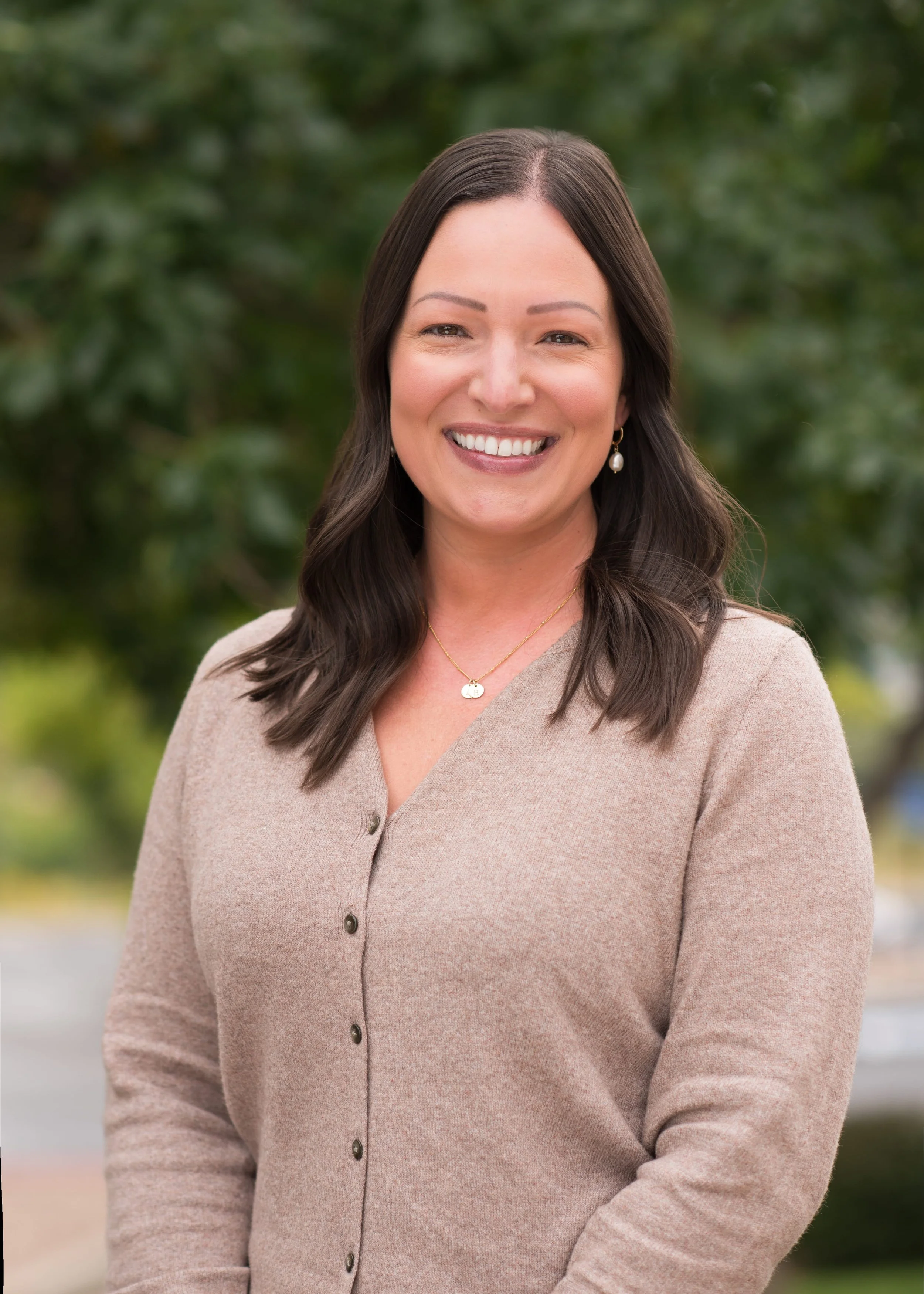 A woman smiling outdoors with blurred green foliage background.