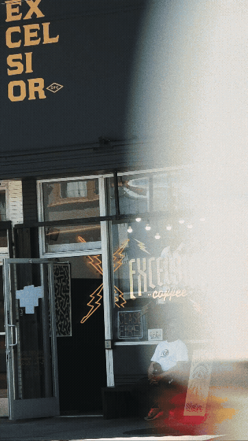 Exterior of a coffee shop with the word "EXCITER" on the sign, and neon lightning bolt decorations visible through the window, with some reflections and a person sitting outside.
