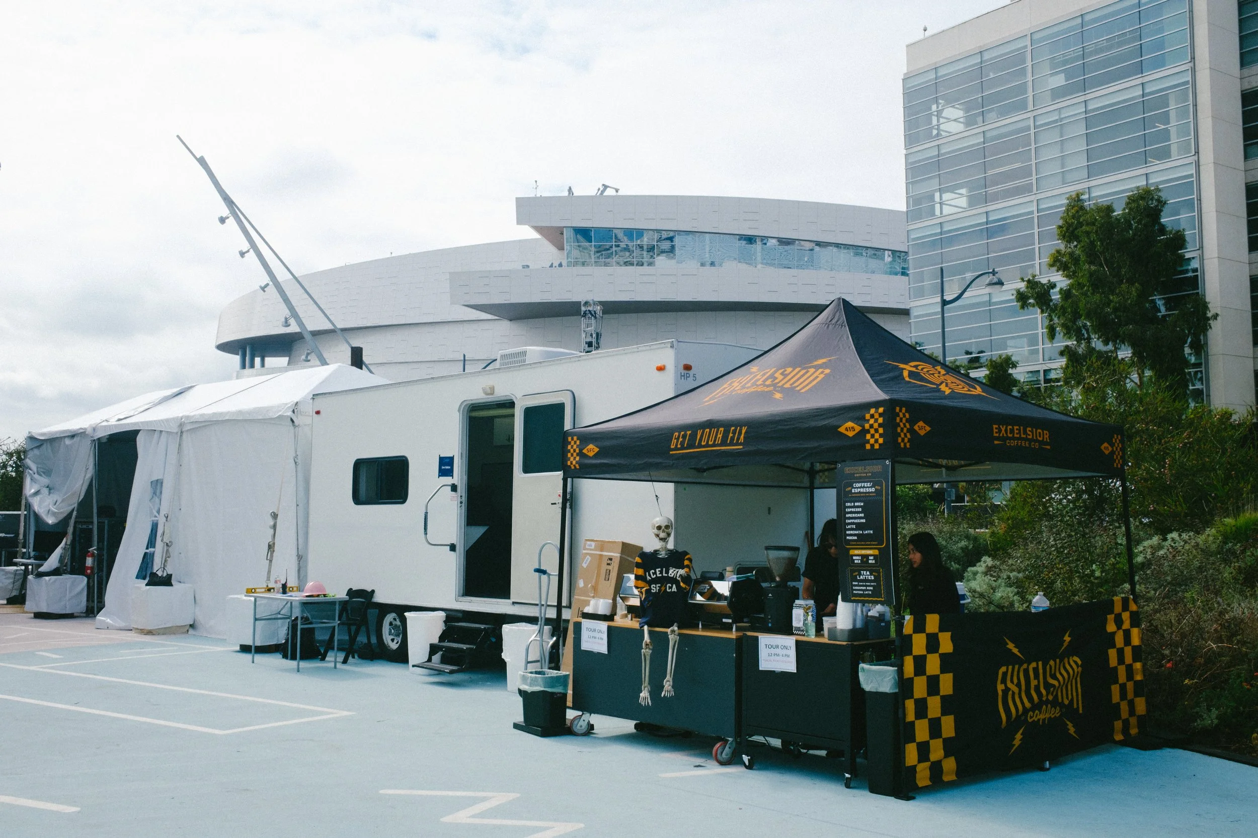 Food truck with a black canopy labeled Excelsior Coffee Co. serving coffee, with a skeletal skeleton waiter and a skeleton. The food truck is outside in a parking lot next to a large modern building, greenery, and cloudy sky.