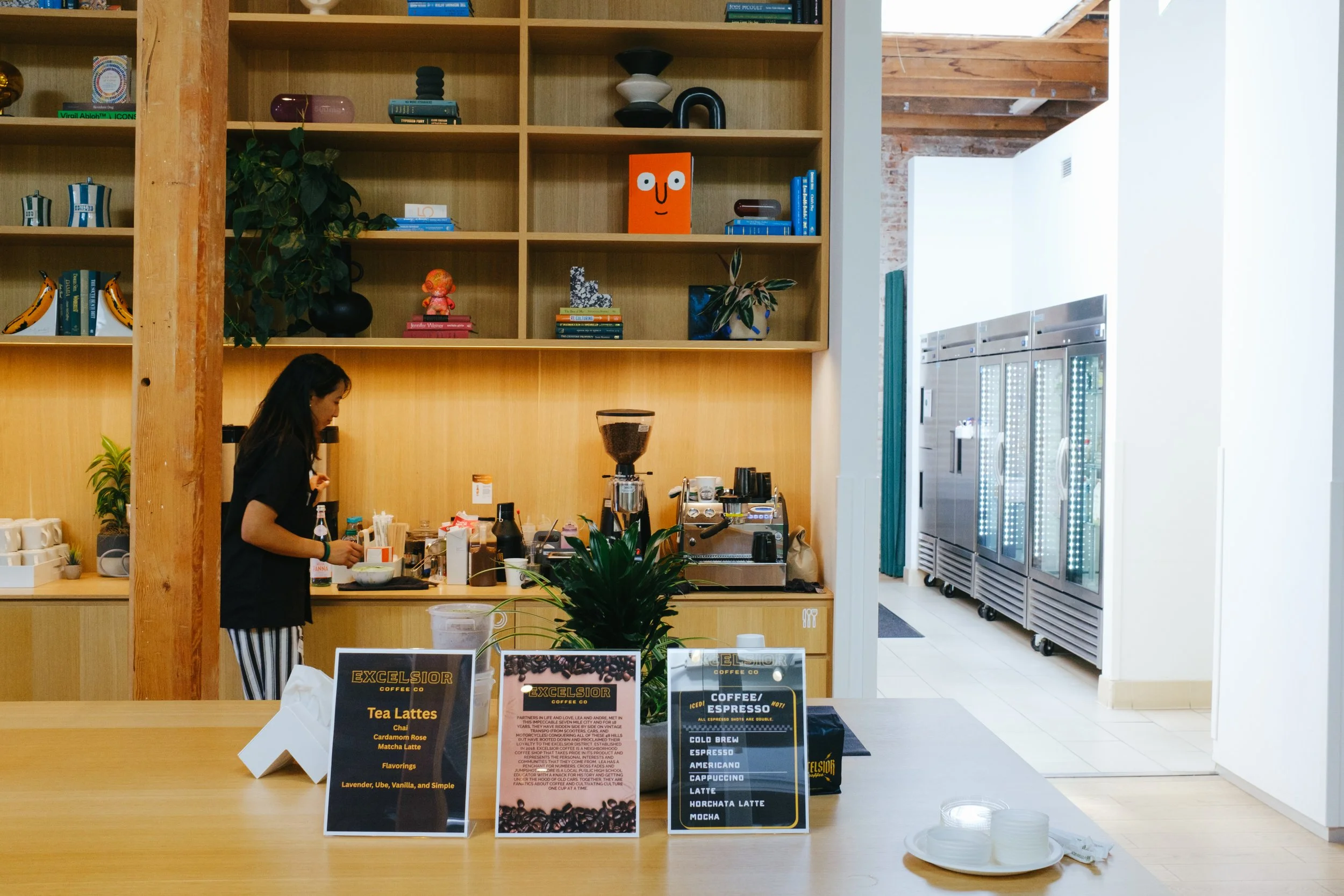 Interior of a modern coffee shop with a barista preparing drinks behind the counter, wooden shelves with decorative objects and books, and refrigerated display cases with beverages in the background.