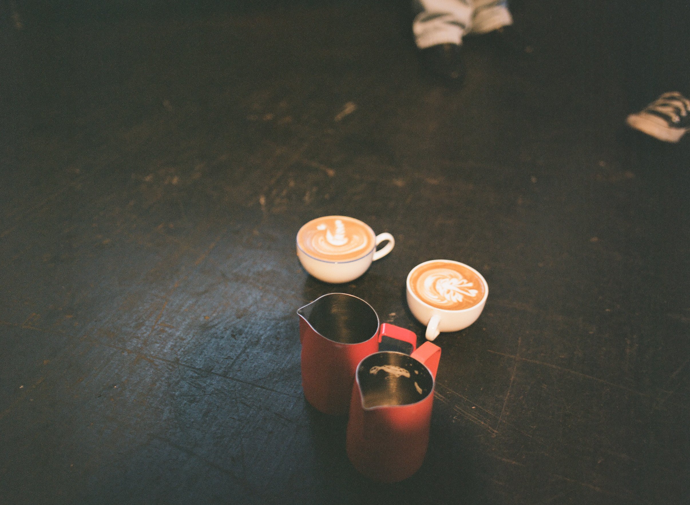 Two cups of coffee with latte art on a dark wooden table, alongside a small red pitcher and a smaller black container.