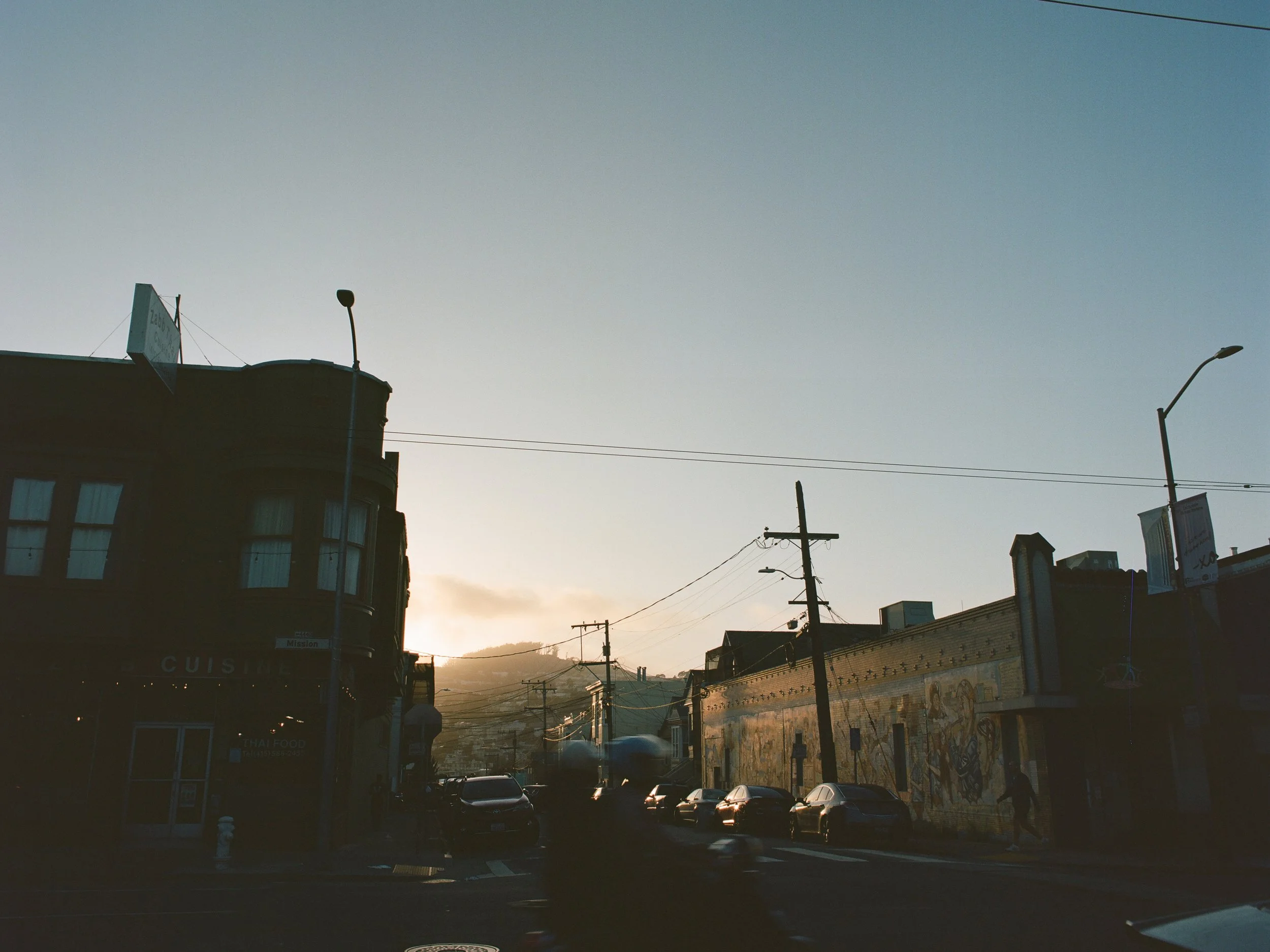 Street scene at sunset with parked cars, utility poles, a building with mural art, and a hill in the background.