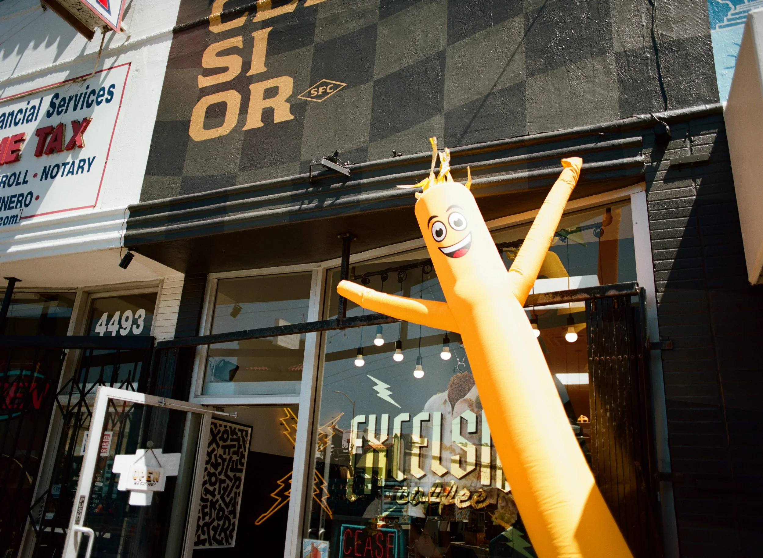 An inflatable happy yellow balloon character with a smiling face and one eye winking, attached to a black building exterior with a glass storefront, during daytime.