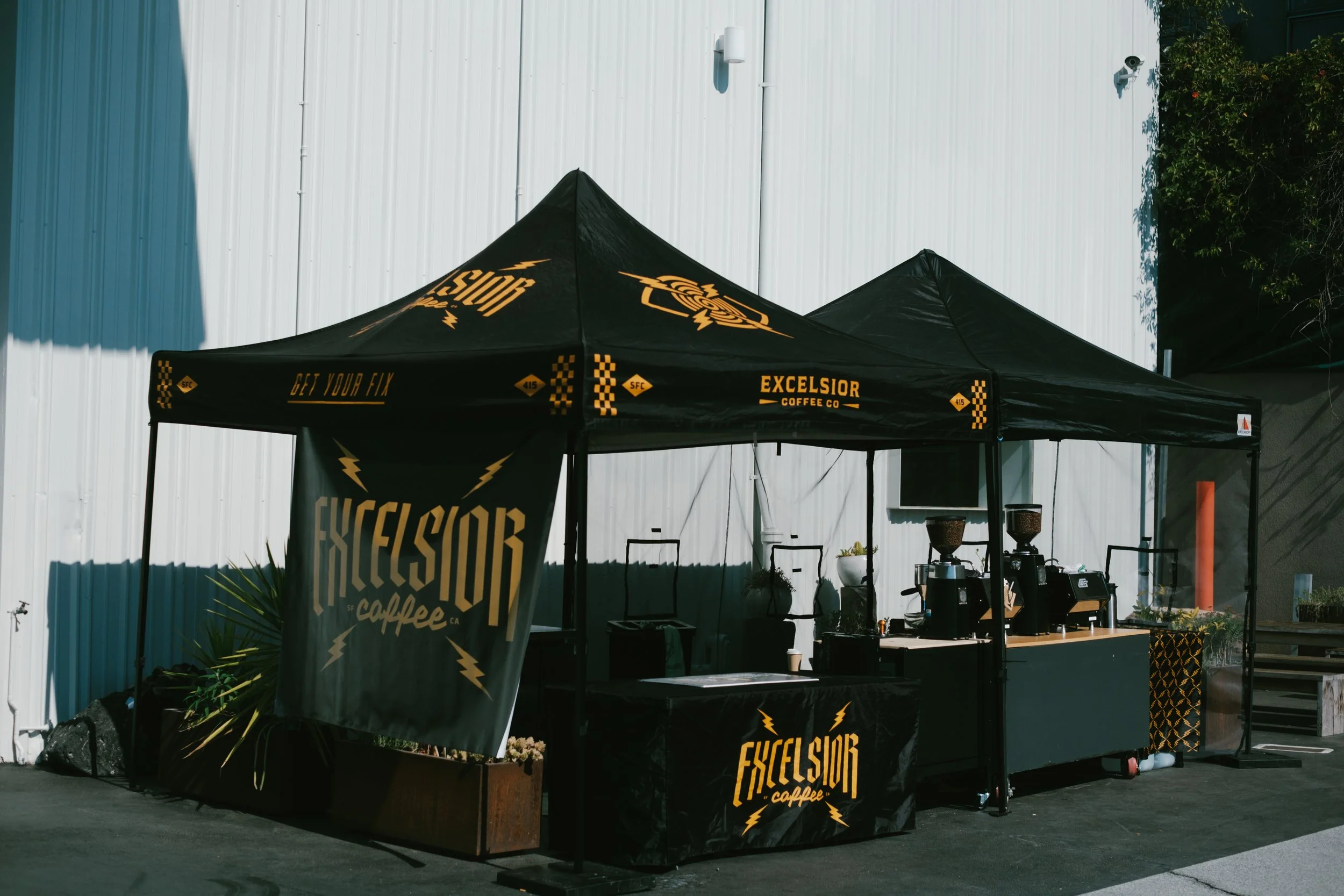 Outdoor coffee stand with black and yellow branding signage reading 'Excelsior Coffee Co.' and 'Get Your Fix,' set up under a black canopy tent with branding, against a white metal wall, with some small plants at the base and coffee equipment visible on the counter.